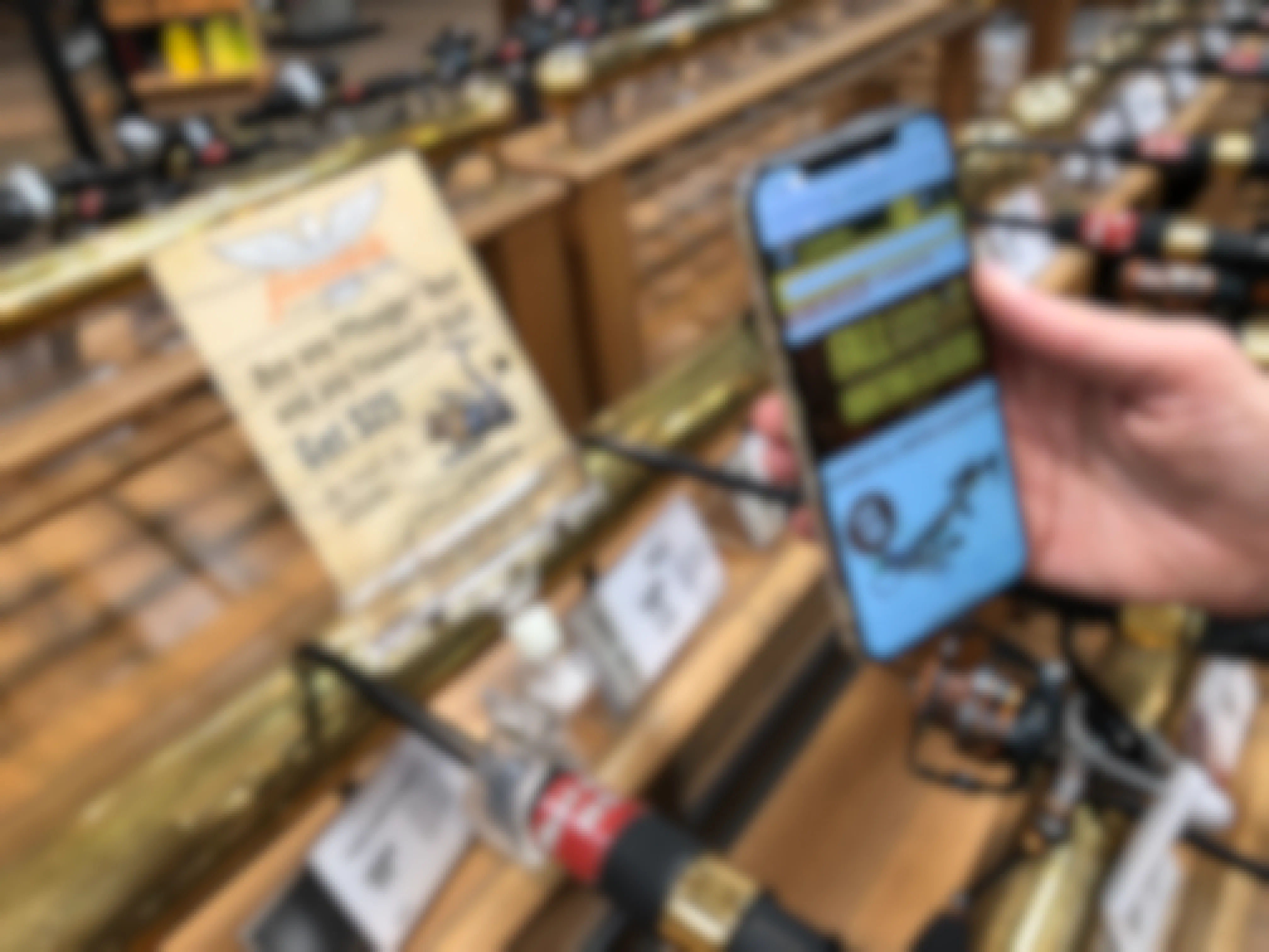 A person's hand holding their cellphone, with the Cabela's site open, in front of a display of fishing rods.