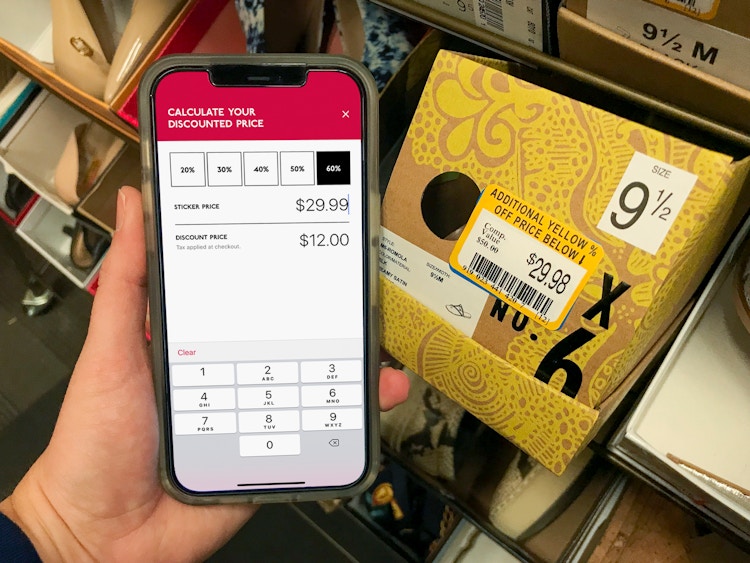 A person holding and iPhone next to a shoe box with a yellow clearance sticker on it. The cell phone screen displays the Discount Calculator inside the DSW app.