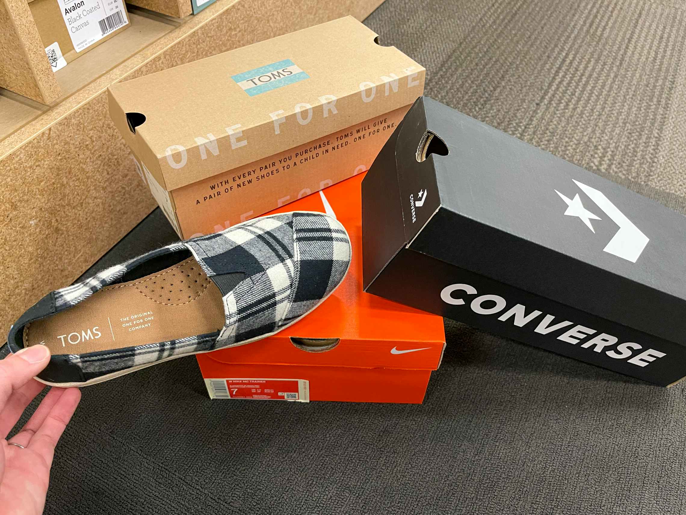 Converse, Nike, and TOMS shoe boxes stacked up on the floor with a person holding one of the TOMS shoes next to the boxes.