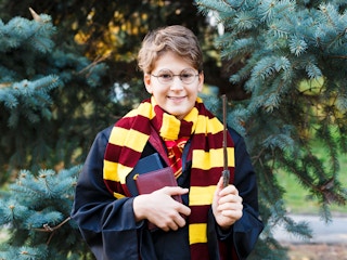 A kid dressed as Harry Potter standing outside and holding a wand and books.