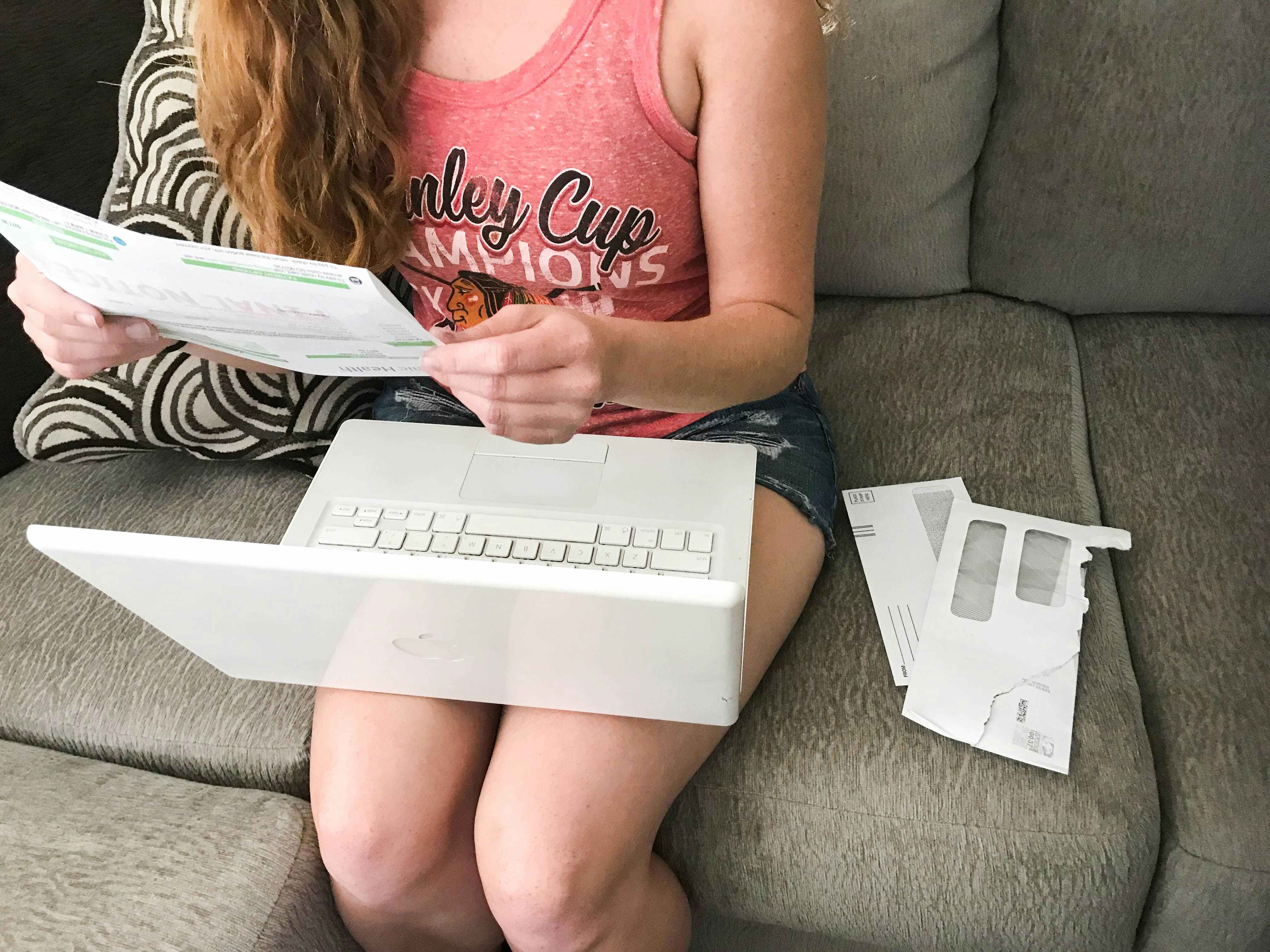 woman from neck down in a pink shirt holds bills with laptop on her lap