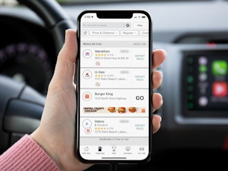 A person sitting in their car, holding up a cell phone displaying the Gas Buddy app