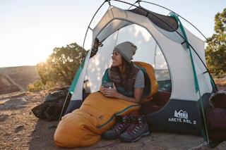 A woman sitting in a sleeping bag, half way out of an open tent. The tent is set up on a mountain top and the sun is rising in the distance.