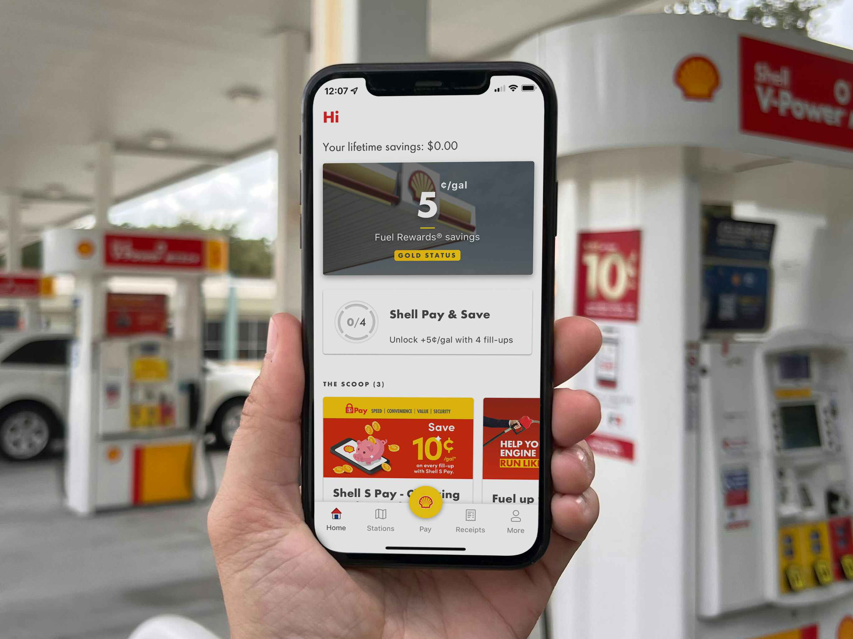 A person's hand holding up a cell phone displaying the Shell Rewards app in front of a Shell gas station