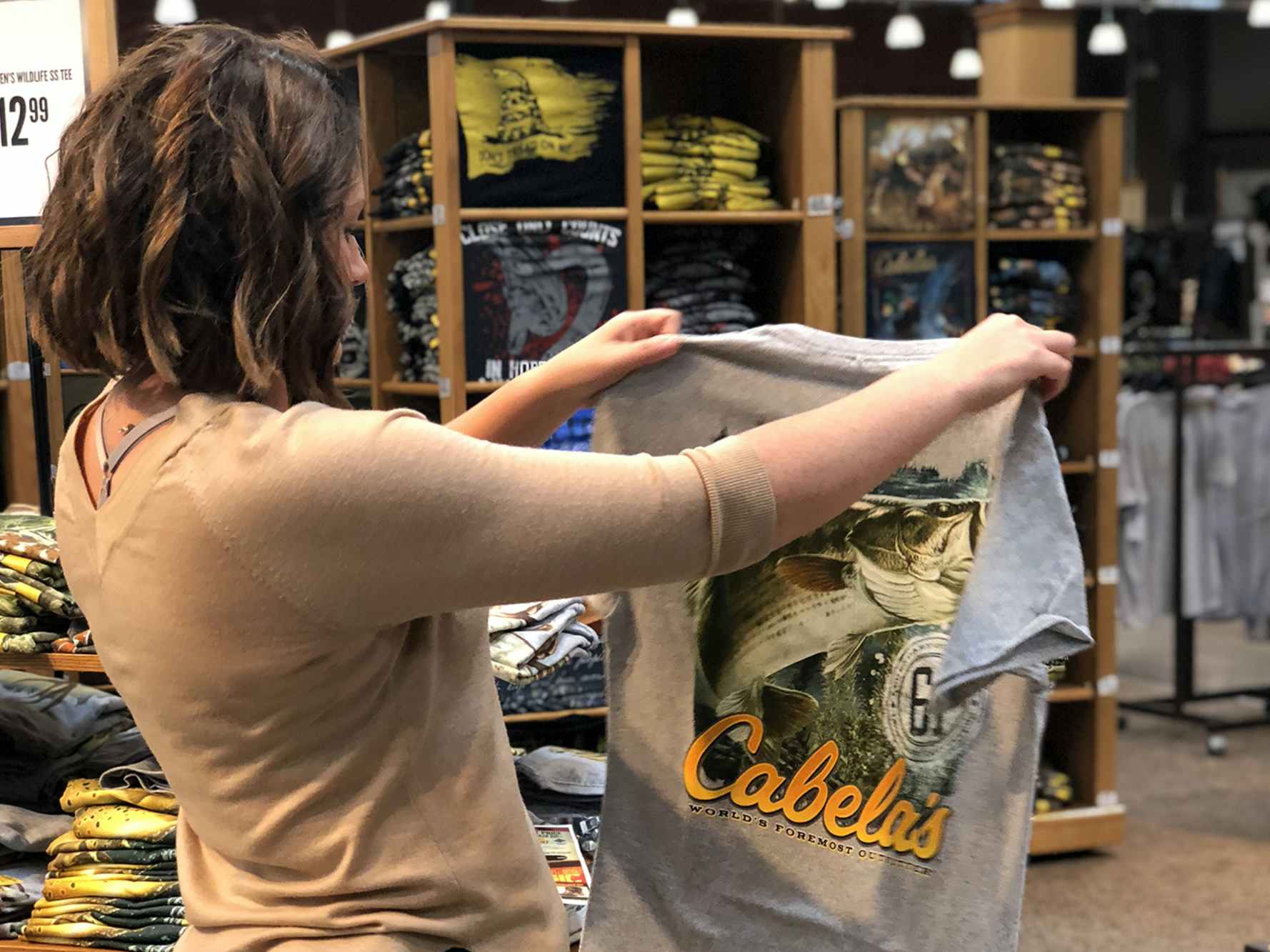 A woman holding up and looking at a Cabela's t-shirt in the clothing section of Cabela's.