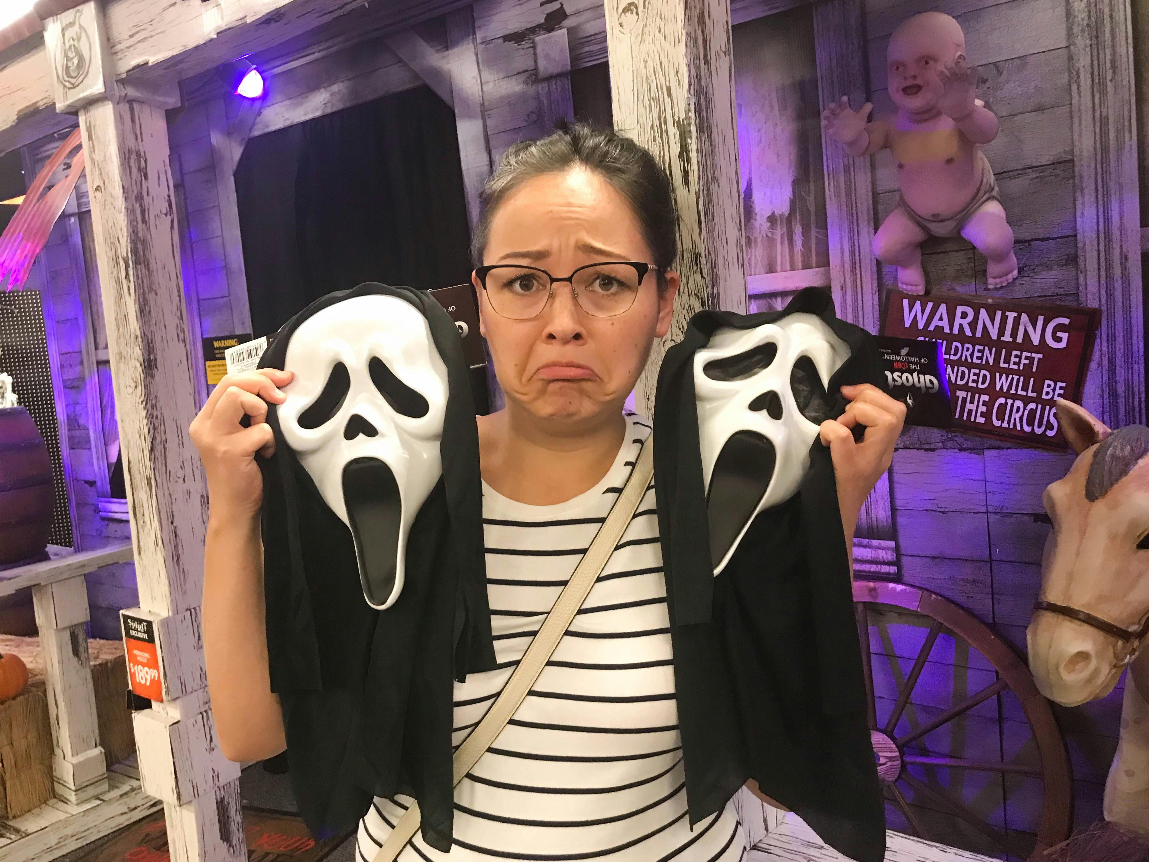 A woman holding up two ghostface masks and frowning