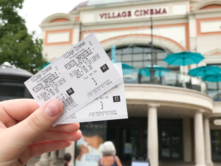 A person's hand holding two movie tickets for Mission Impossible in front of a theater showing the sales price of $5.30.