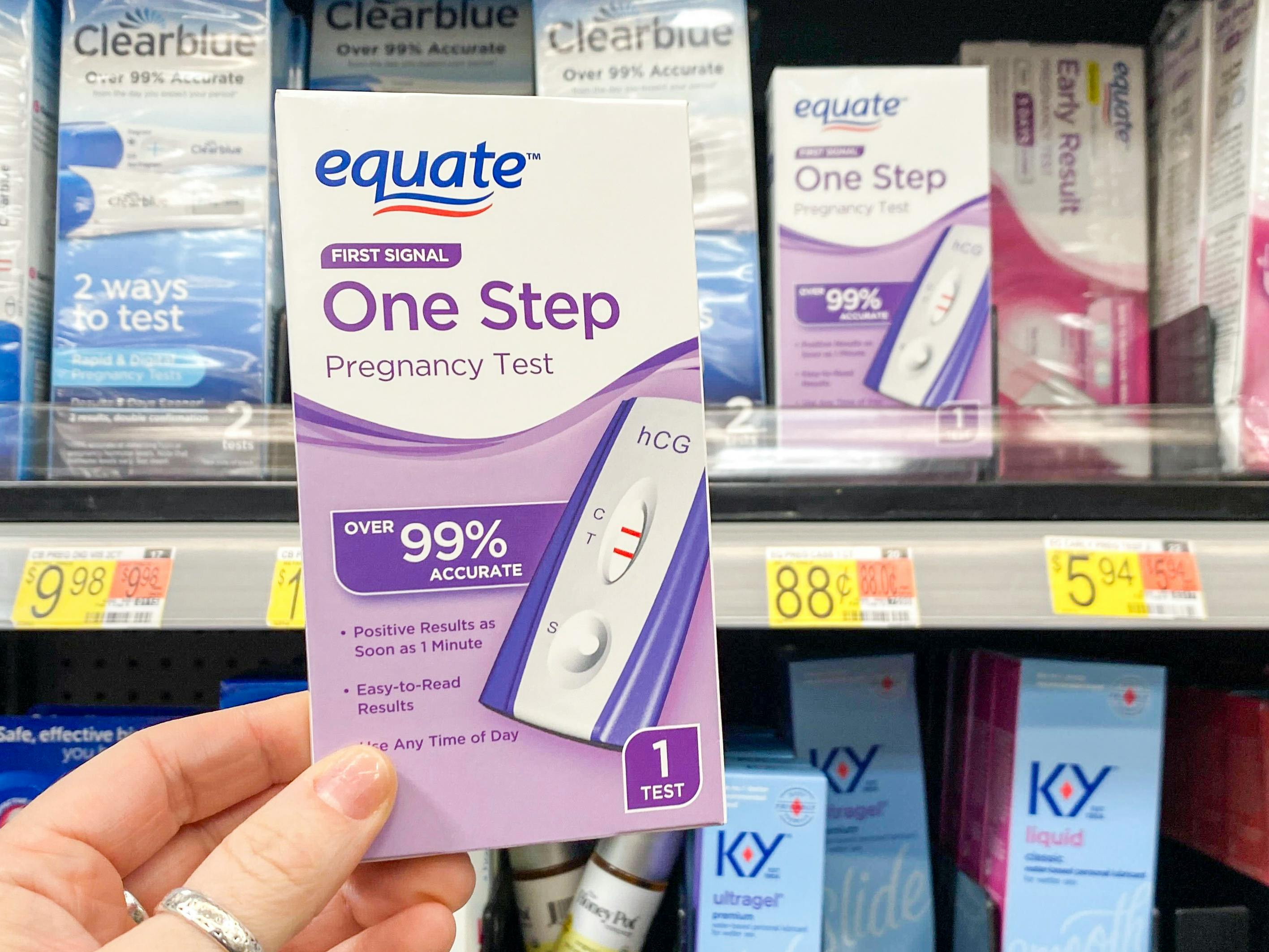 A person holding an Equate pregnancy test in front of the pregnancy test shelf at Walmart.