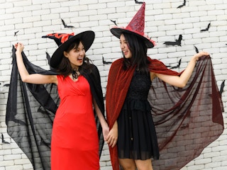Two women dressed as witches smiling at each other in front of a white brick wall.