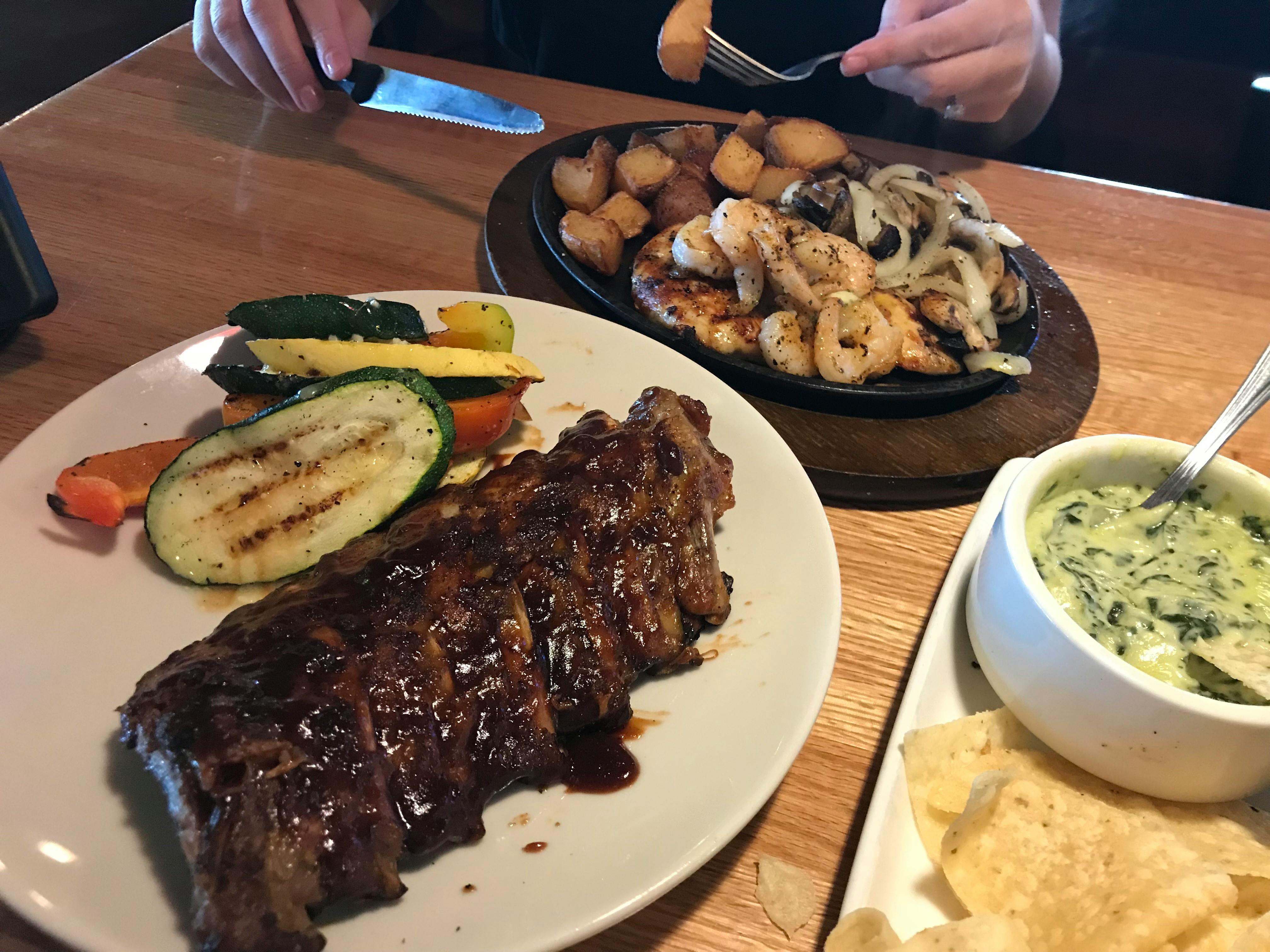 A plate of ribs and vegetables on a table next to a skillet plate of shrimp, chicken, potatoes, and onions, and another plate with a bowl of spinach-artichoke dip and tortilla chips on a table at Applebee's.