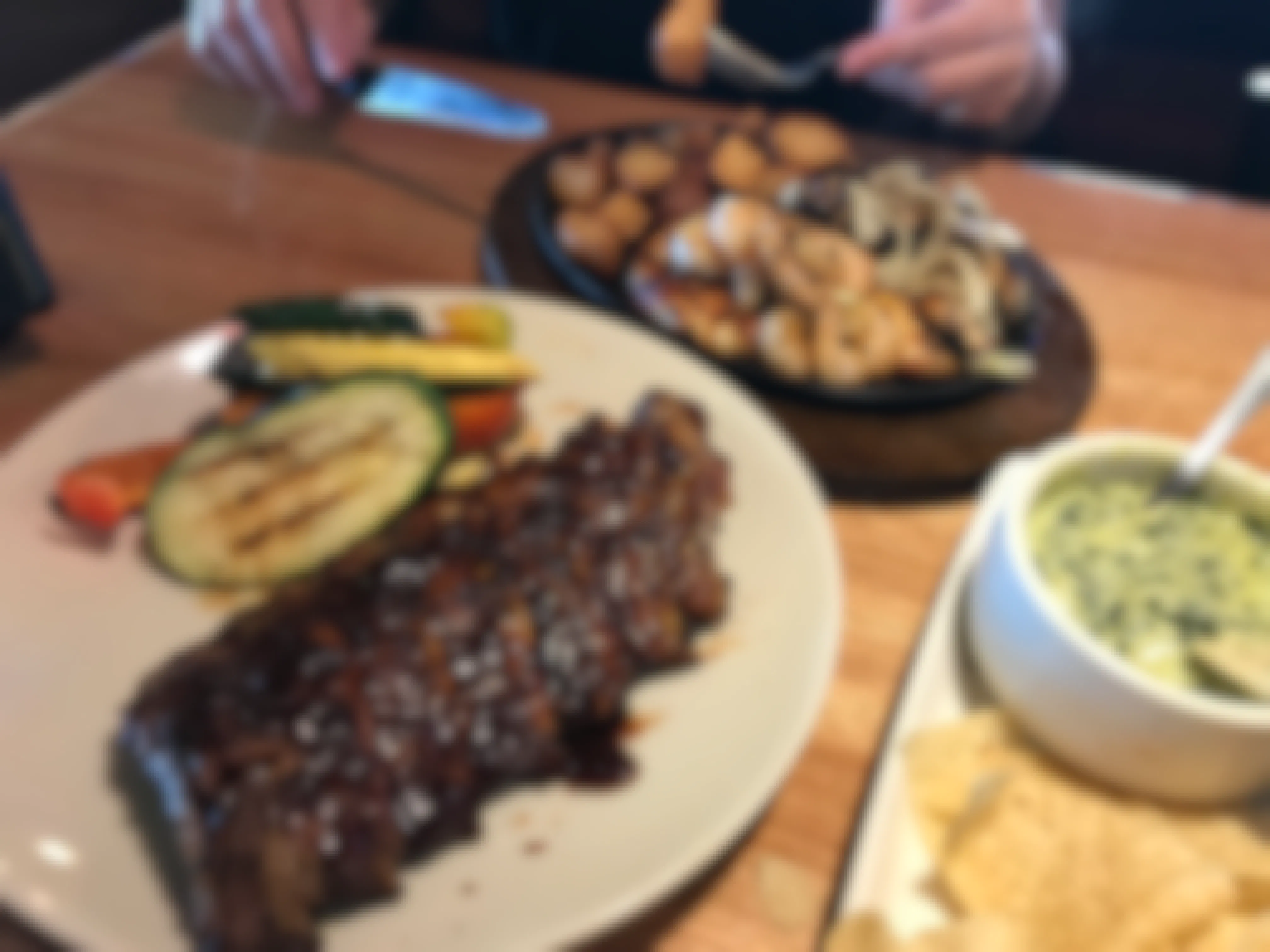 A plate of ribs and vegetables on a table next to a skillet plate of shrimp, chicken, potatoes, and onions, and another plate with a bowl of spinach-artichoke dip and tortilla chips on a table at Applebee's.