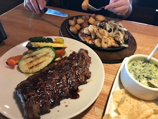 A plate of ribs and vegetables on a table next to a skillet plate of shrimp, chicken, potatoes, and onions, and another plate with a bowl of spinach-artichoke dip and tortilla chips on a table at Applebee's.