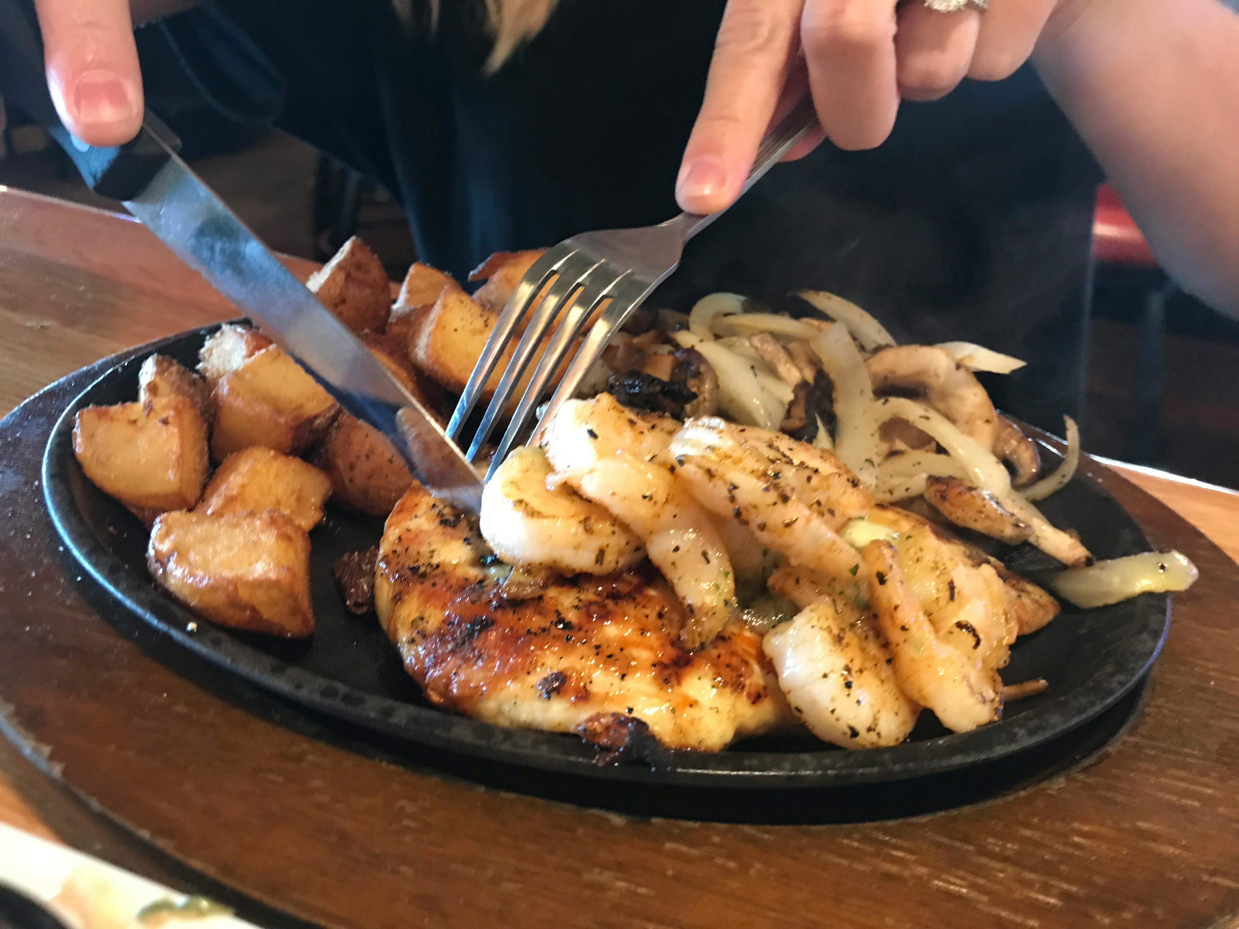 A person's hands using a fork and knife to cut into some chicken on a skillet plate of chicken, shrimp, potatoes, and onions, on a table at Applebee's.