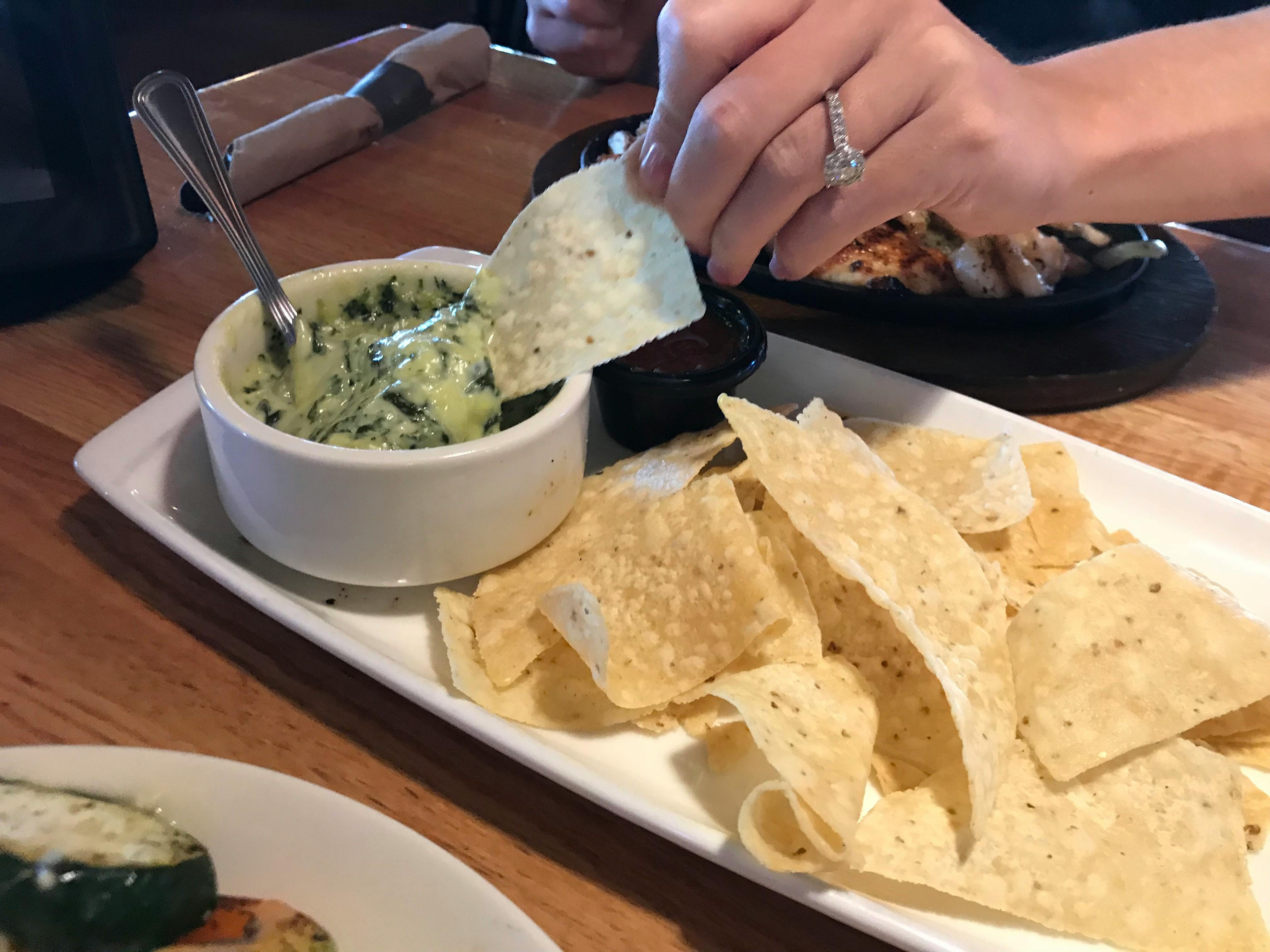 A person's hand dipping a tortilla chip into a bowl of Applebee's Spinach Artichoke dip next to more tortilla chips on a platter.