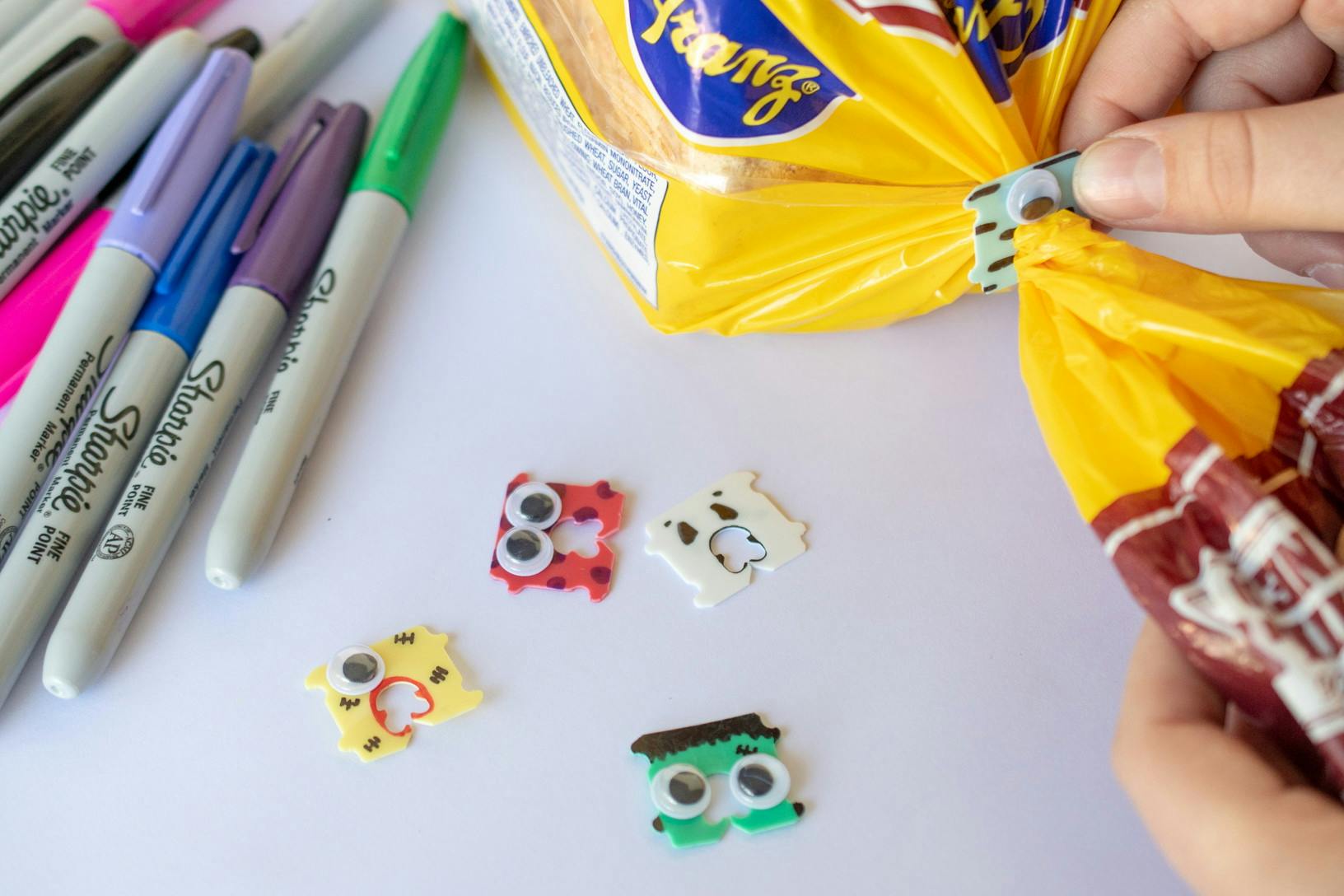 Bread clips decorated like Halloween monsters next to varying colors of sharpie pens and a person attaching one bread clip to a loaf of bread