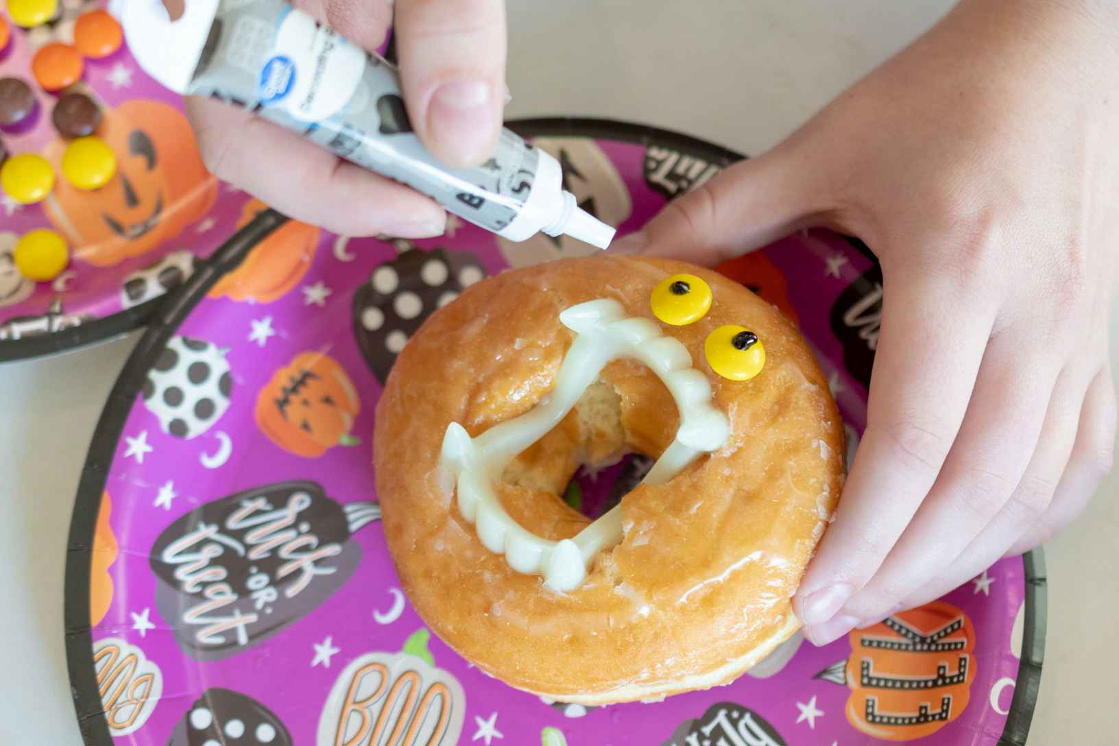 A person using black icing to add dots to the center of Reese's pieces candies attached to a donut. Plastic vampire teeth are inserted into the center of the donut.