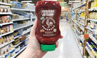 A person's hand holding up a bottle of Sriracha ketchup in an aisle of a grocery store.