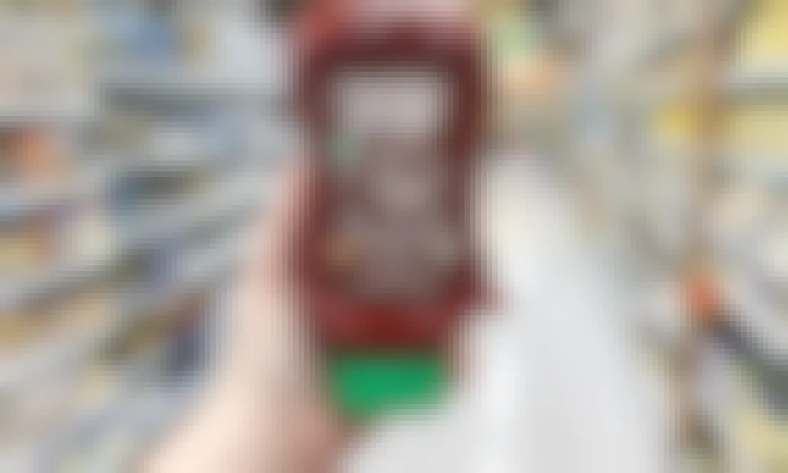 A person's hand holding up a bottle of Sriracha ketchup in an aisle of a grocery store.