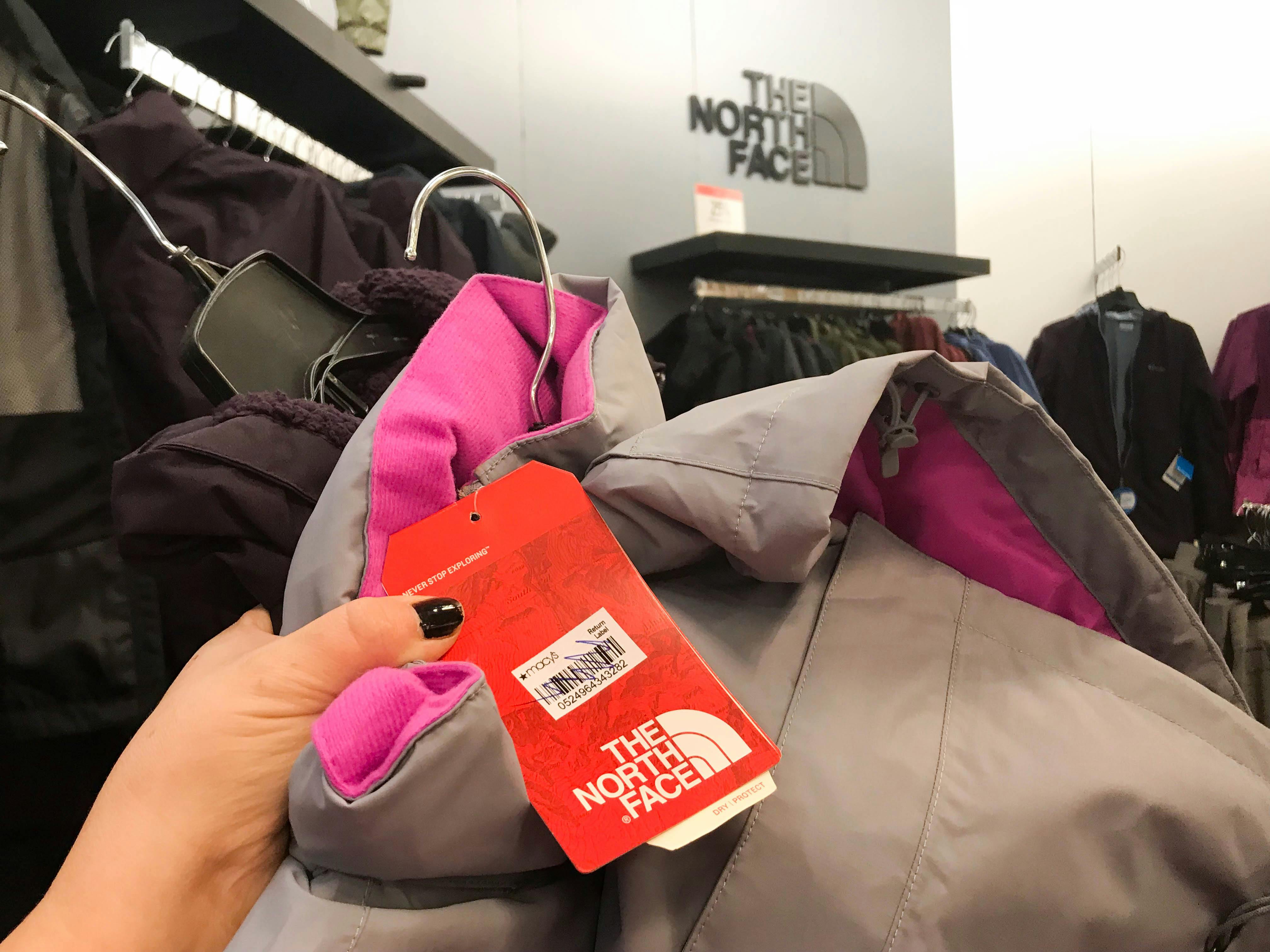 A person's hand taking a The North Face coat and showing the tag at Macy's.