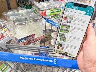 A person holding a cellphone displaying Ball canning jars on the Amazon app to compare prices with Walmart products sitting in a shopping cart behind the phone.