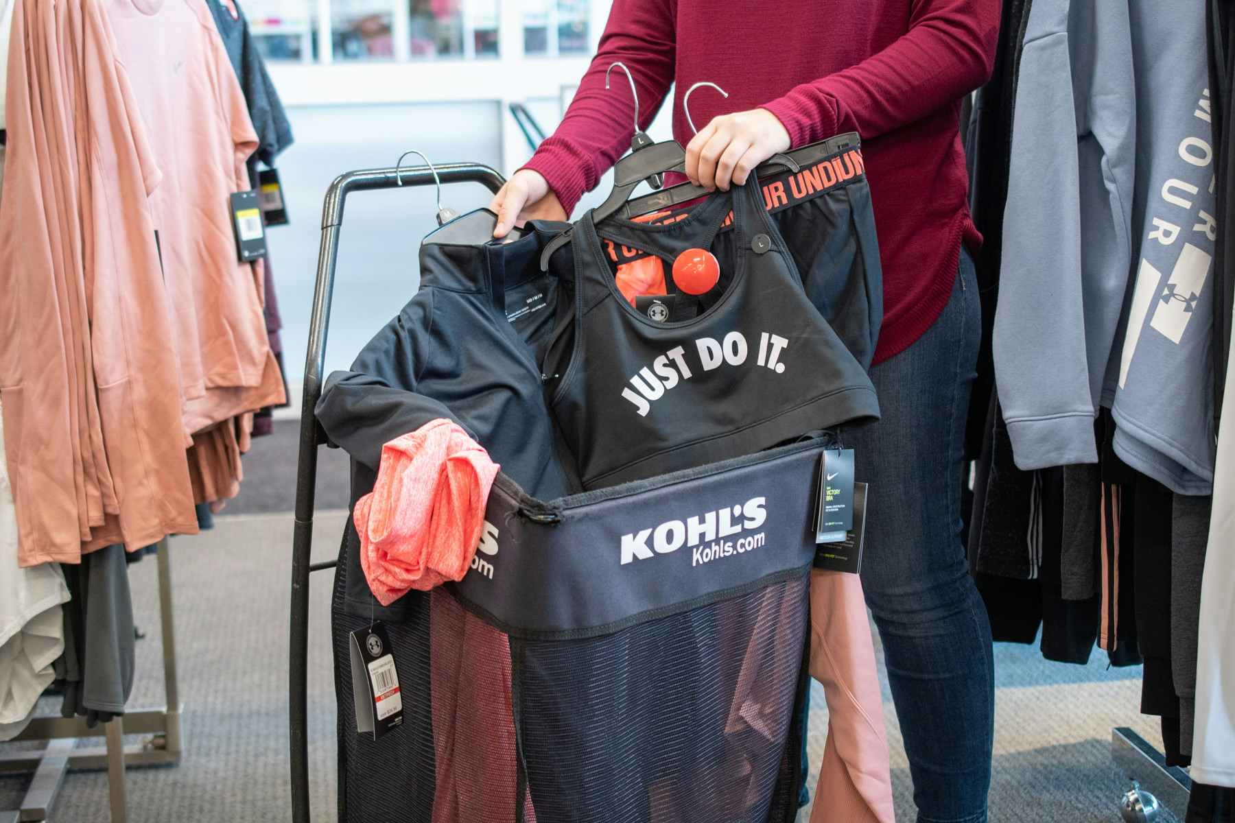 A person putting Nike and Under Armour activewear into a Kohl's shopping cart.
