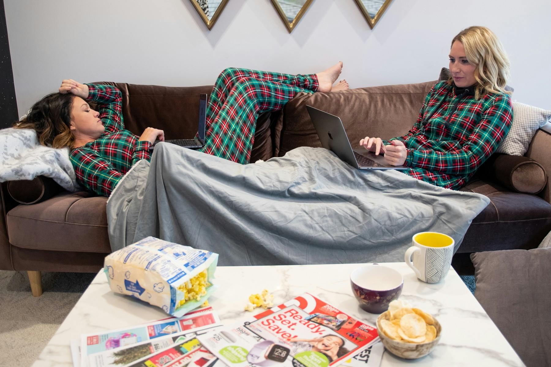 Two women in matching pajamas lounging on a couch, both of them using their laptops to shop Black Friday online deals. In the foreground there is a coffee table with paper shopping ads scattered on it next to some popcorn and coffee mugs.