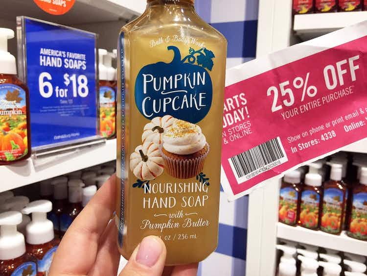 A person's hand holding up a Pumpkin Cupcake scented hand soap and a coupon for 25% off next to a sign that reads "hand soaps 6 for $18" in Bath & Body Works.