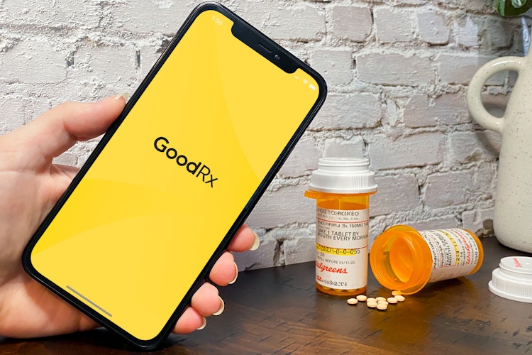 Someone holding up a phone displaying the GoodRX app launch screen with some prescription medication on a table in the background