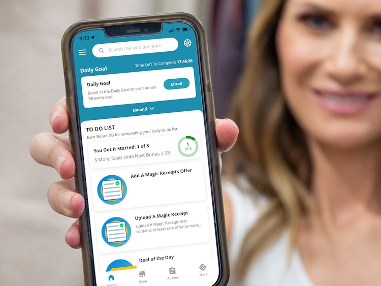 A woman holding up a cell phone displaying the Swagbucks app home screen