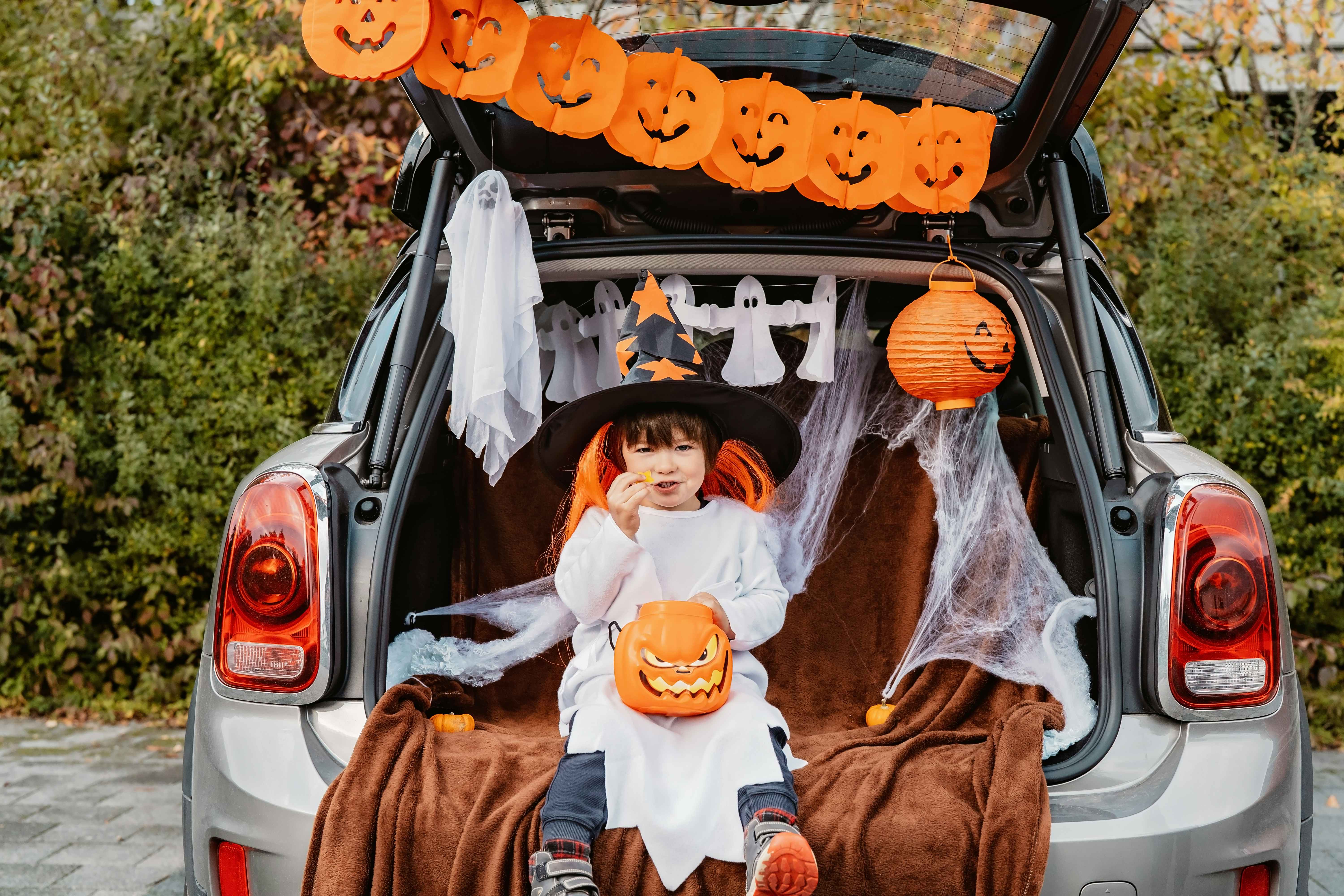 A child dressed in a halloween costume, eating a piece of candy is sitting on in the open truck of a car. The car trunk is decorated with pumpkins and ghosts.