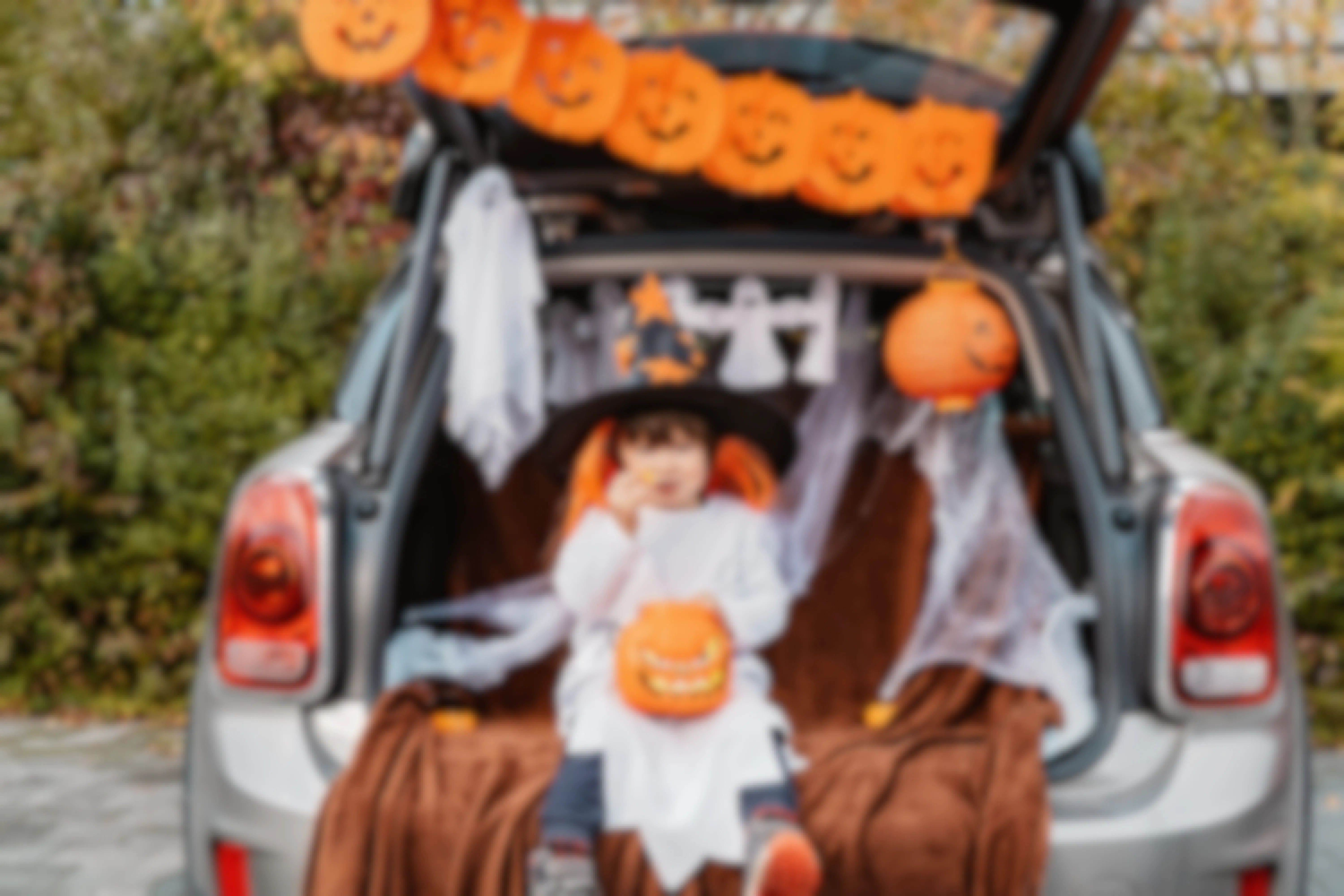 A child dressed in a halloween costume, eating a piece of candy is sitting on in the open truck of a car. The car trunk is decorated with pumpkins and ghosts.
