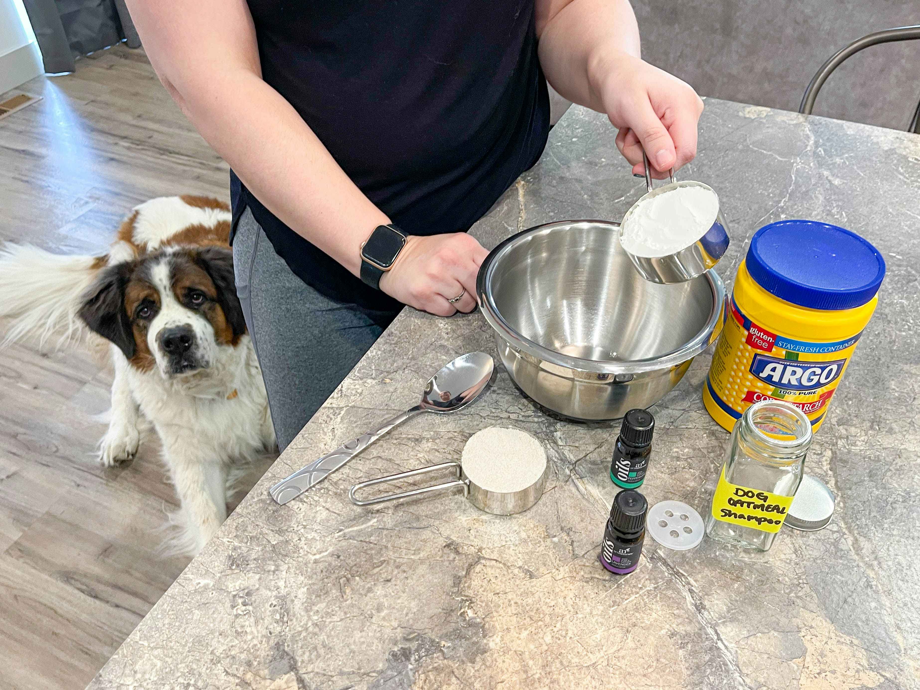 A person putting ingredients for the DIY Dog Oatmeal Shampoo into a metal mixing bowl that is sitting on their kitchen counter next to more of the ingredients. A dog is just behind the person, peering out.