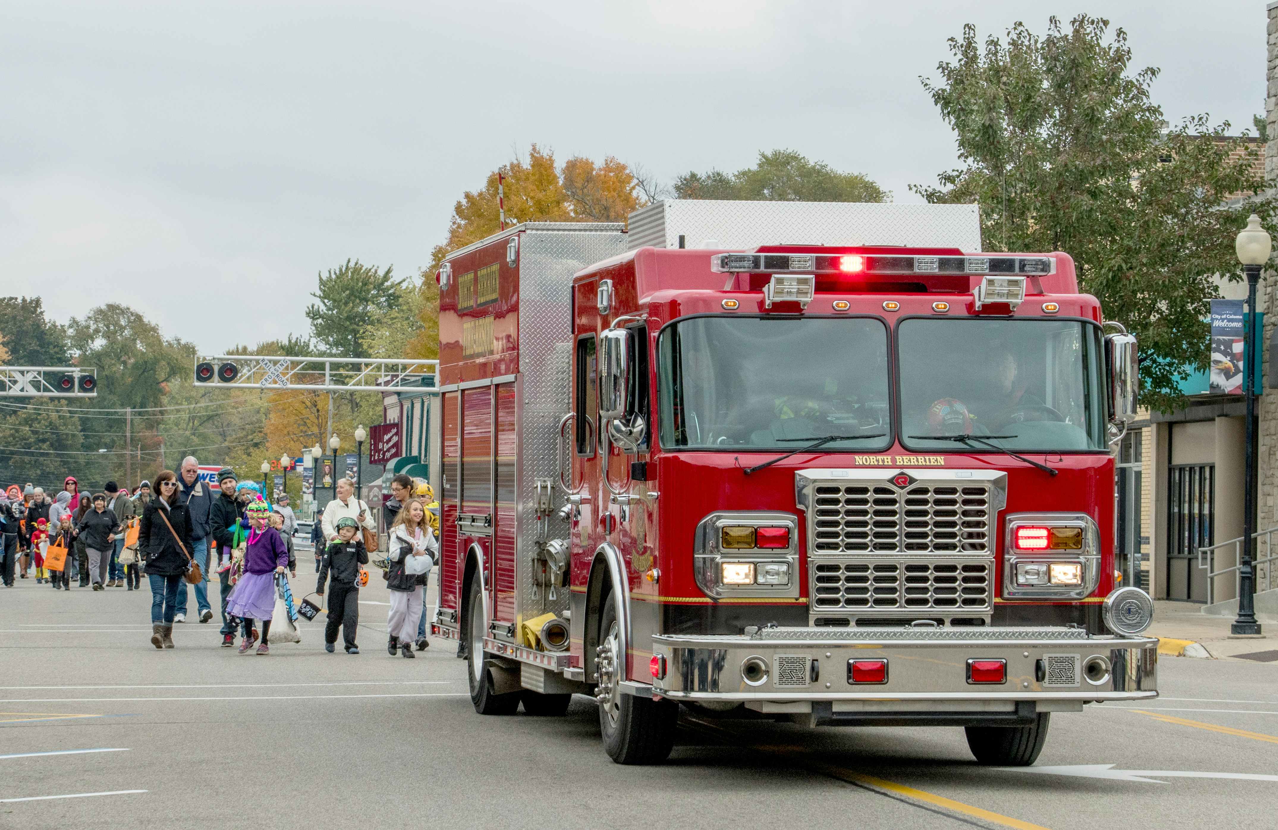 A fire truck driving on a road with adults and kids in halloween costumes walking behind.
