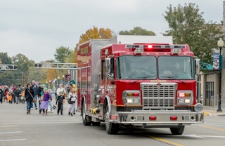 A fire truck driving on a road with adults and kids in halloween costumes walking behind.
