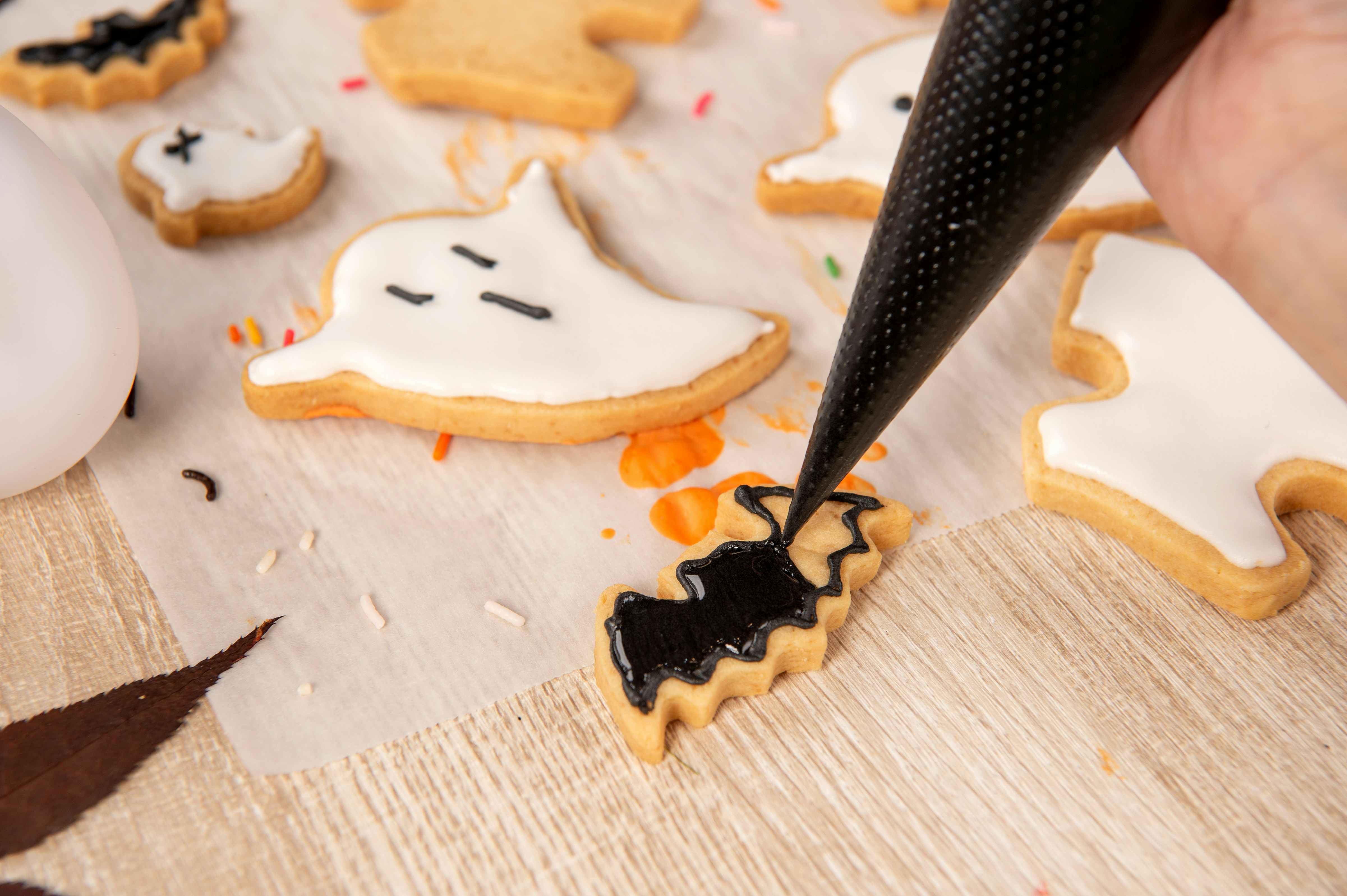 A person piping black icing onto a bat shaped cookie. Other Halloween cookies such as ghosts are laying beside it.