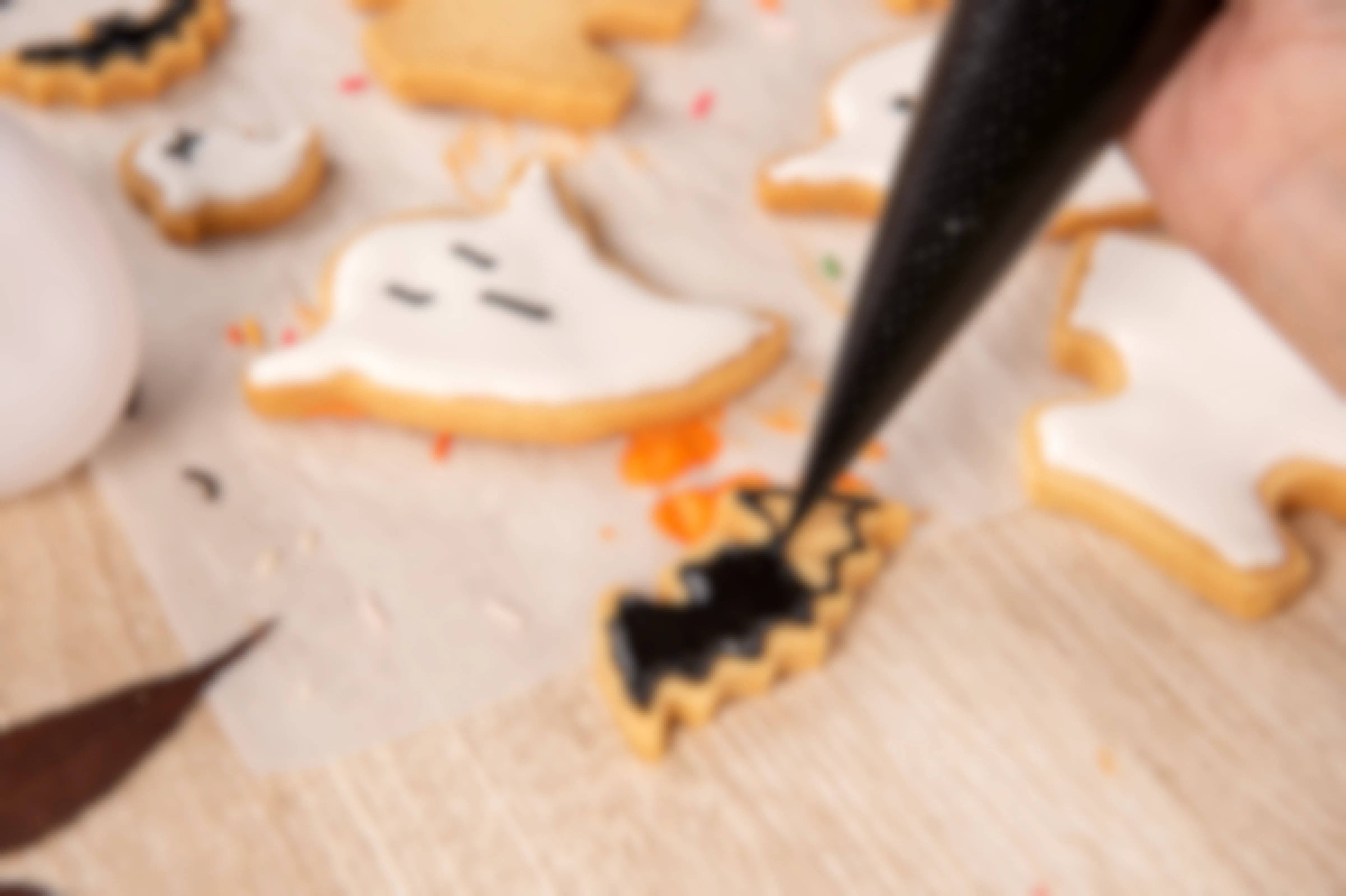A person piping black icing onto a bat shaped cookie. Other Halloween cookies such as ghosts are laying beside it.