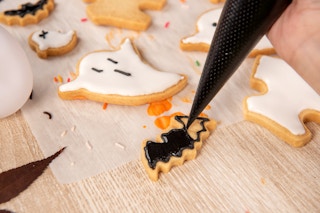 A person piping black icing onto a bat shaped cookie. Other Halloween cookies such as ghosts are laying beside it.