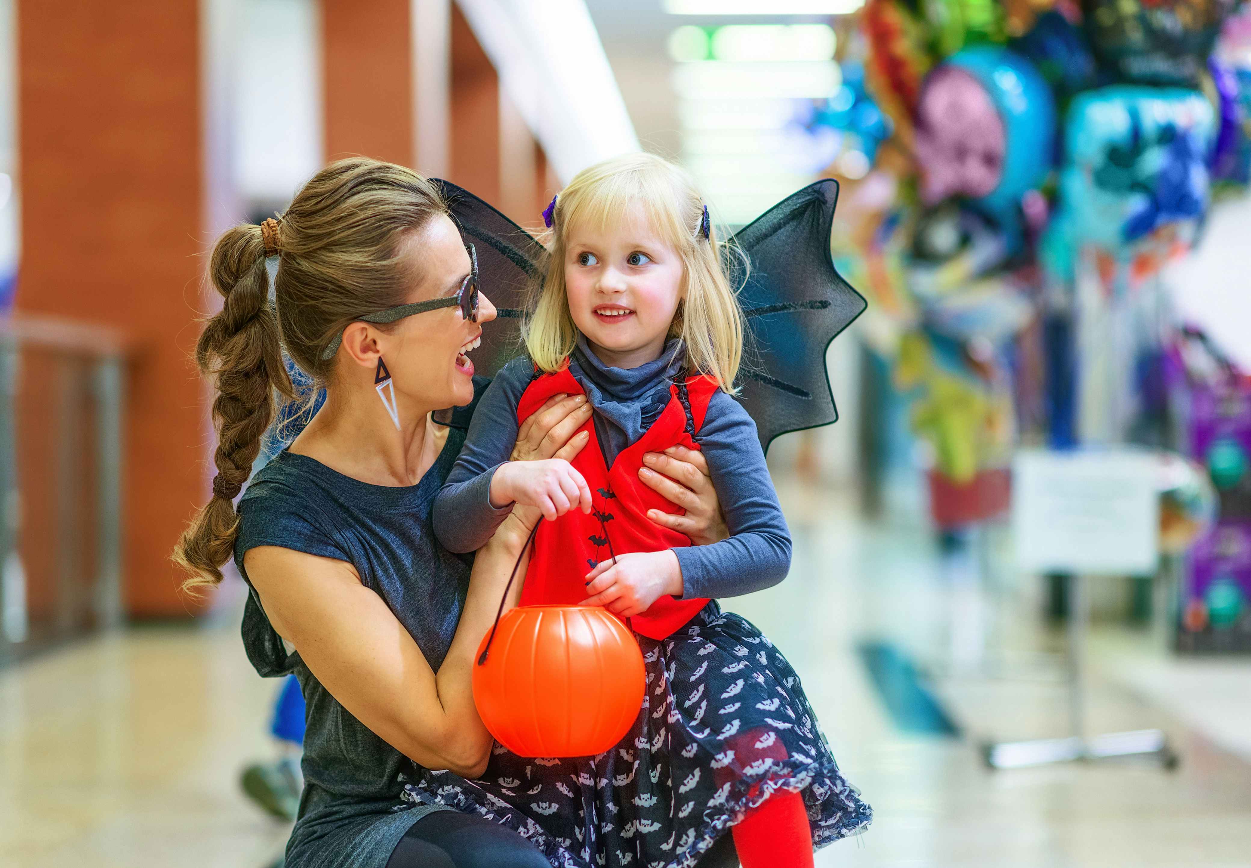 A woman and her daughter dressed in halloween costumes smile at each other, inside a shopping mall.