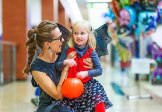 A woman and her daughter dressed in halloween costumes smile at each other, inside a shopping mall.