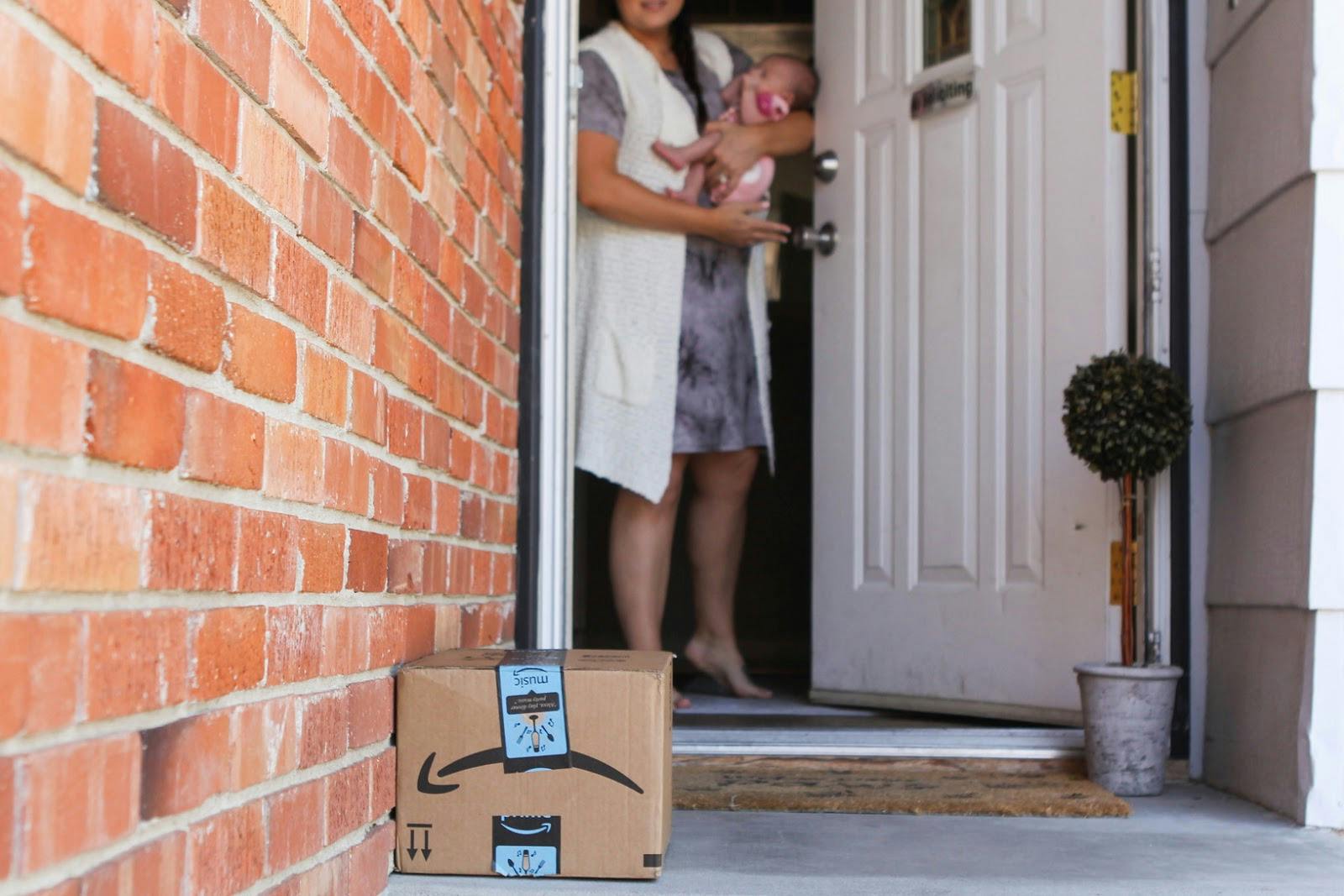 A mom holding a baby, opening the front door to an Amazon delivery box on the porch.