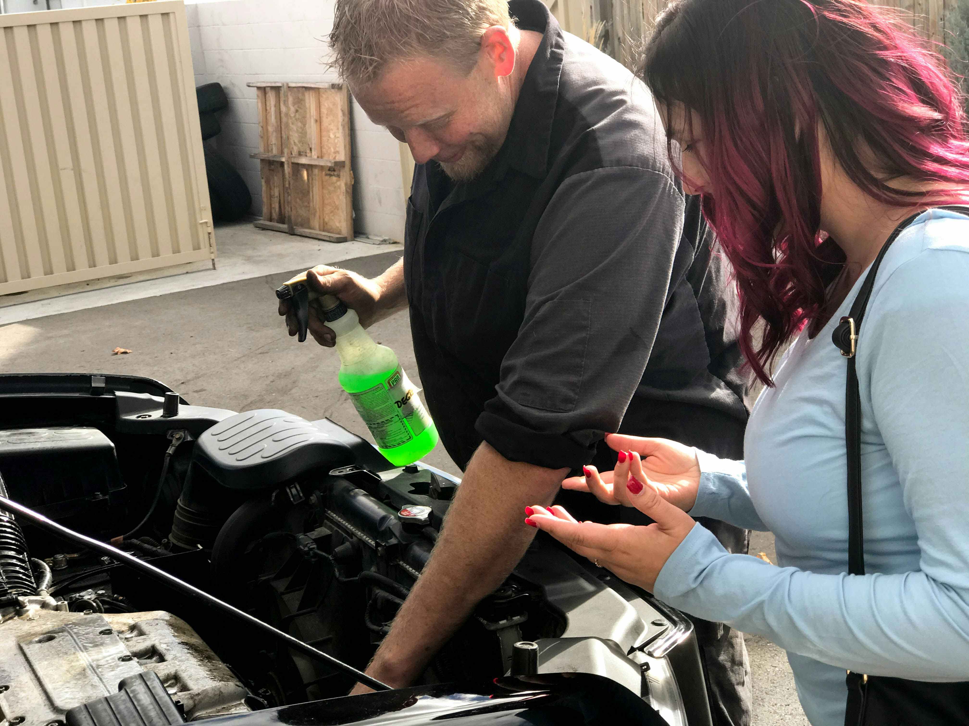 A person talking to an auto shop employee who is working under the hood of a vehicle.