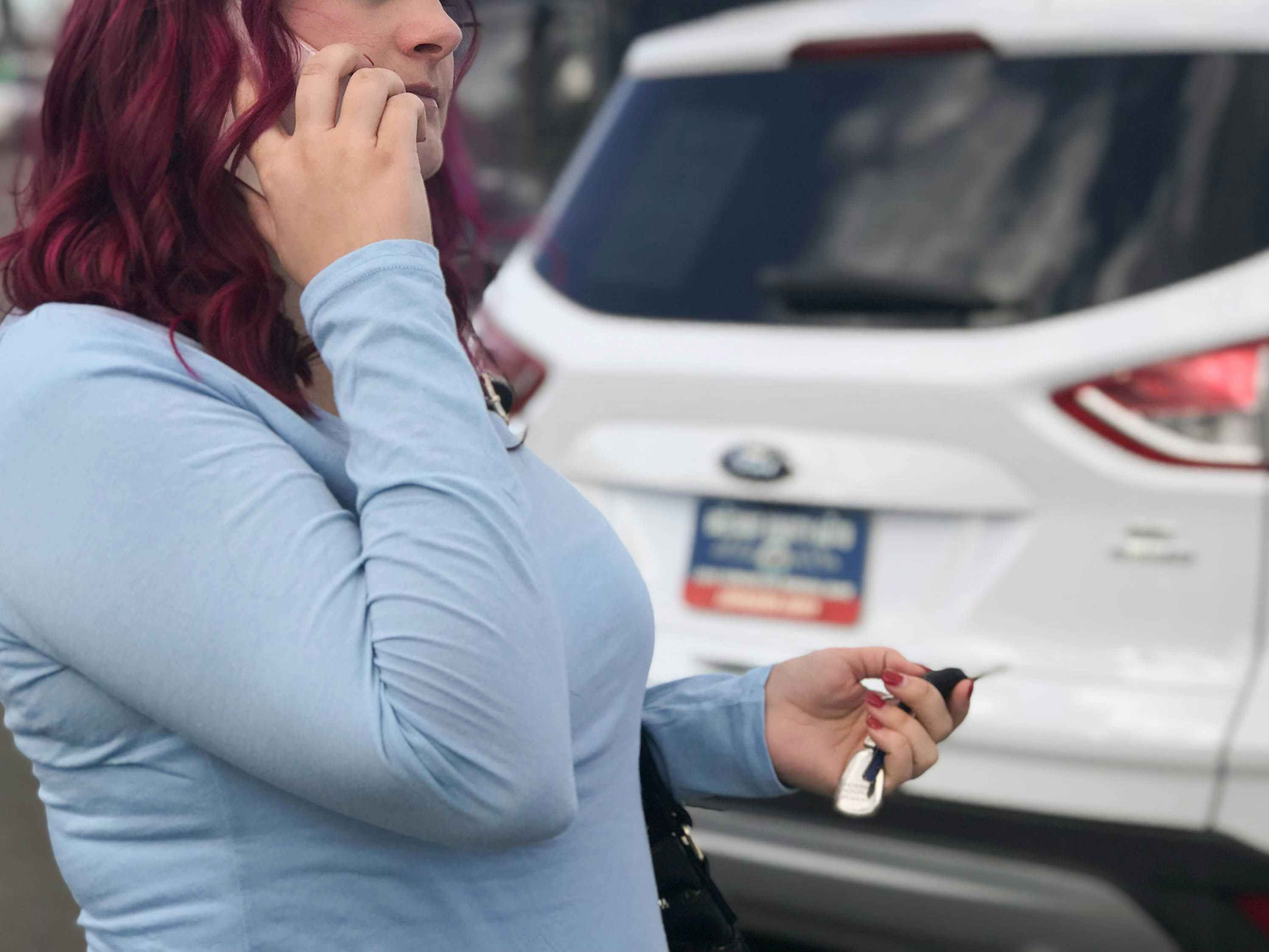 A person on the phone, holding a set of keys and standing in a dealership parking lot.