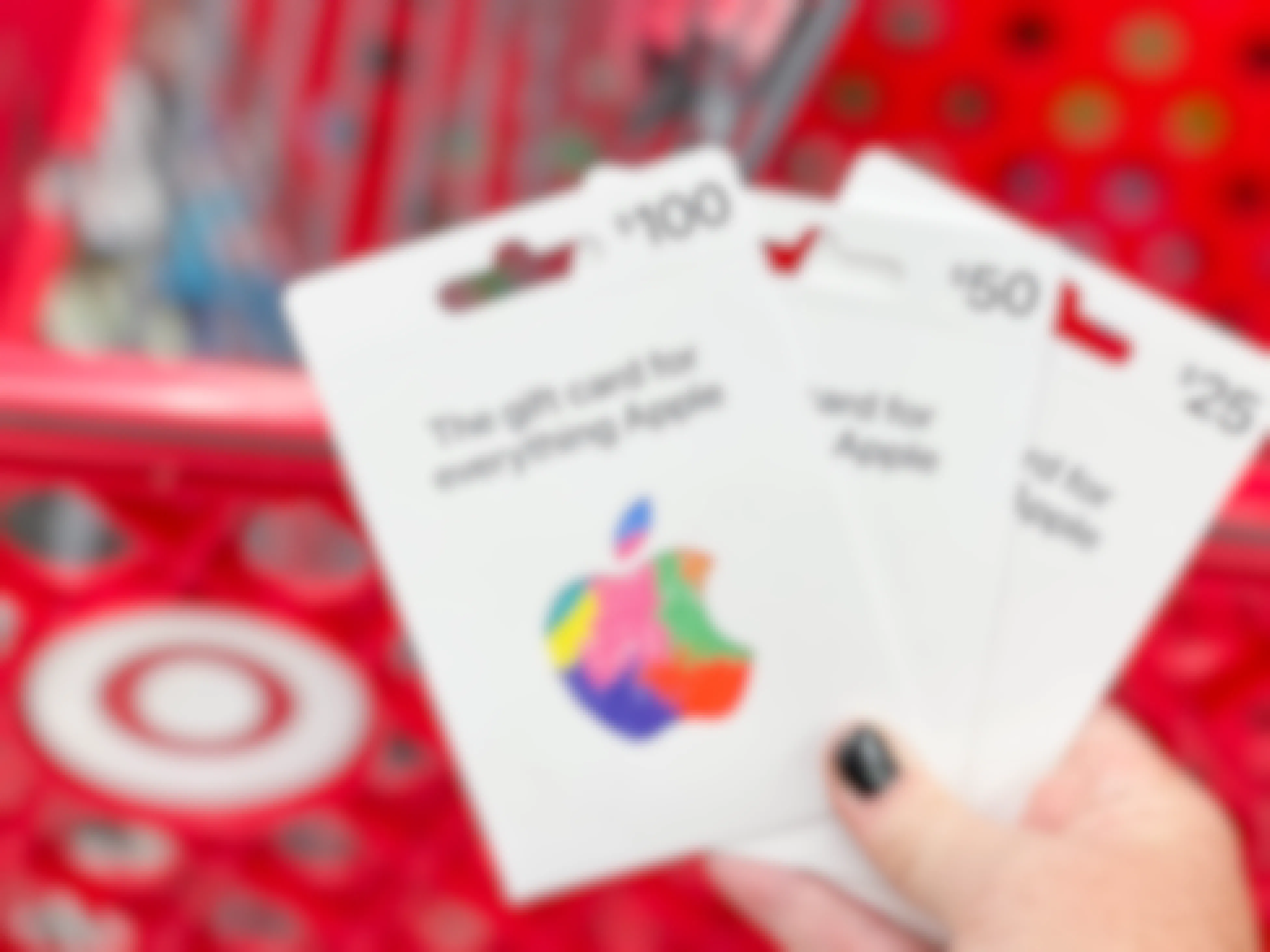 A person's hand holding up three Apple gift cards in front of a Target shopping cart.