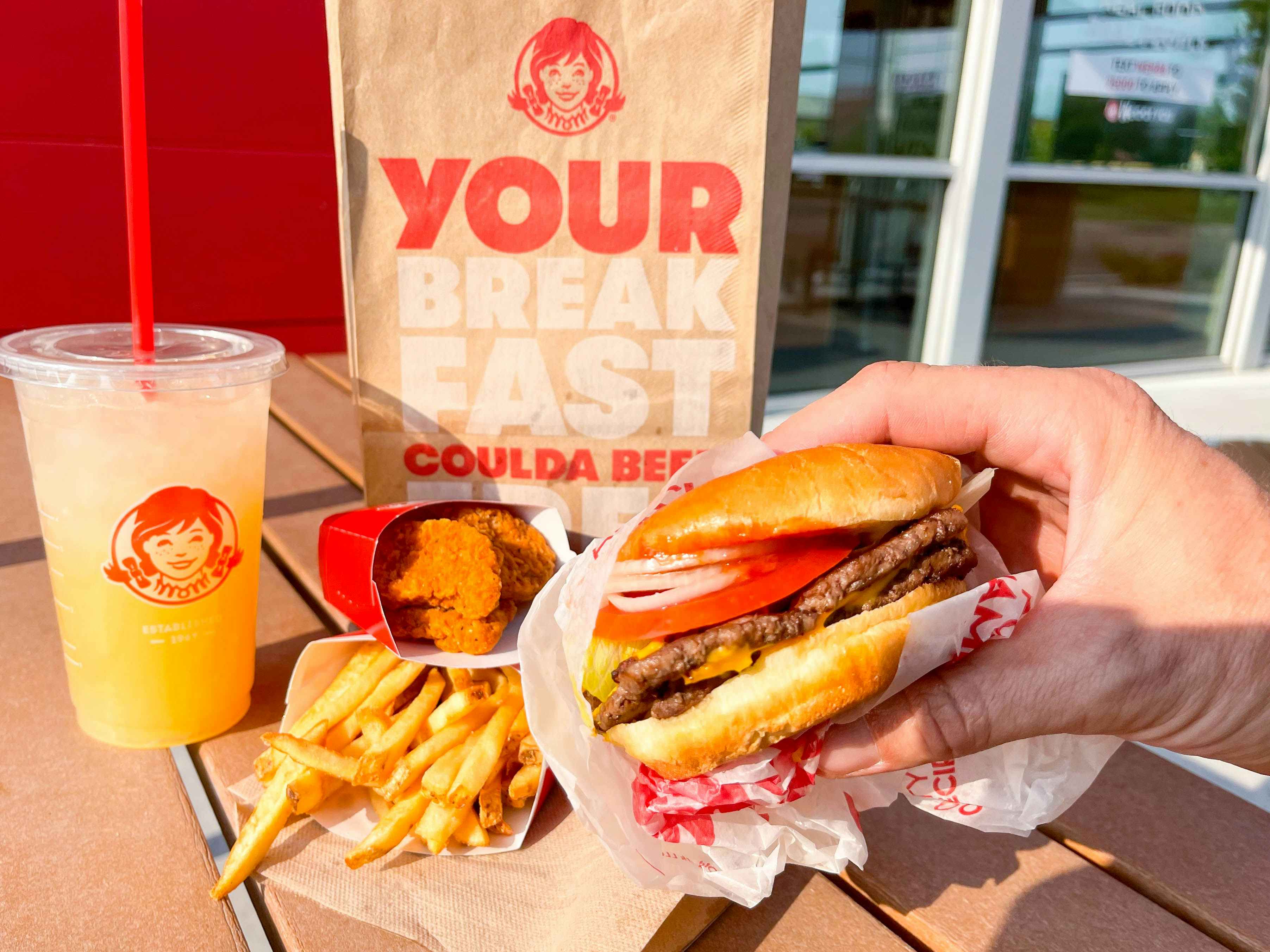 A persons hand holding a Wendy's double stack burger in front of fries, chicken nuggets, a drink, and a Wendy's bag on a table outside of Wendy's.