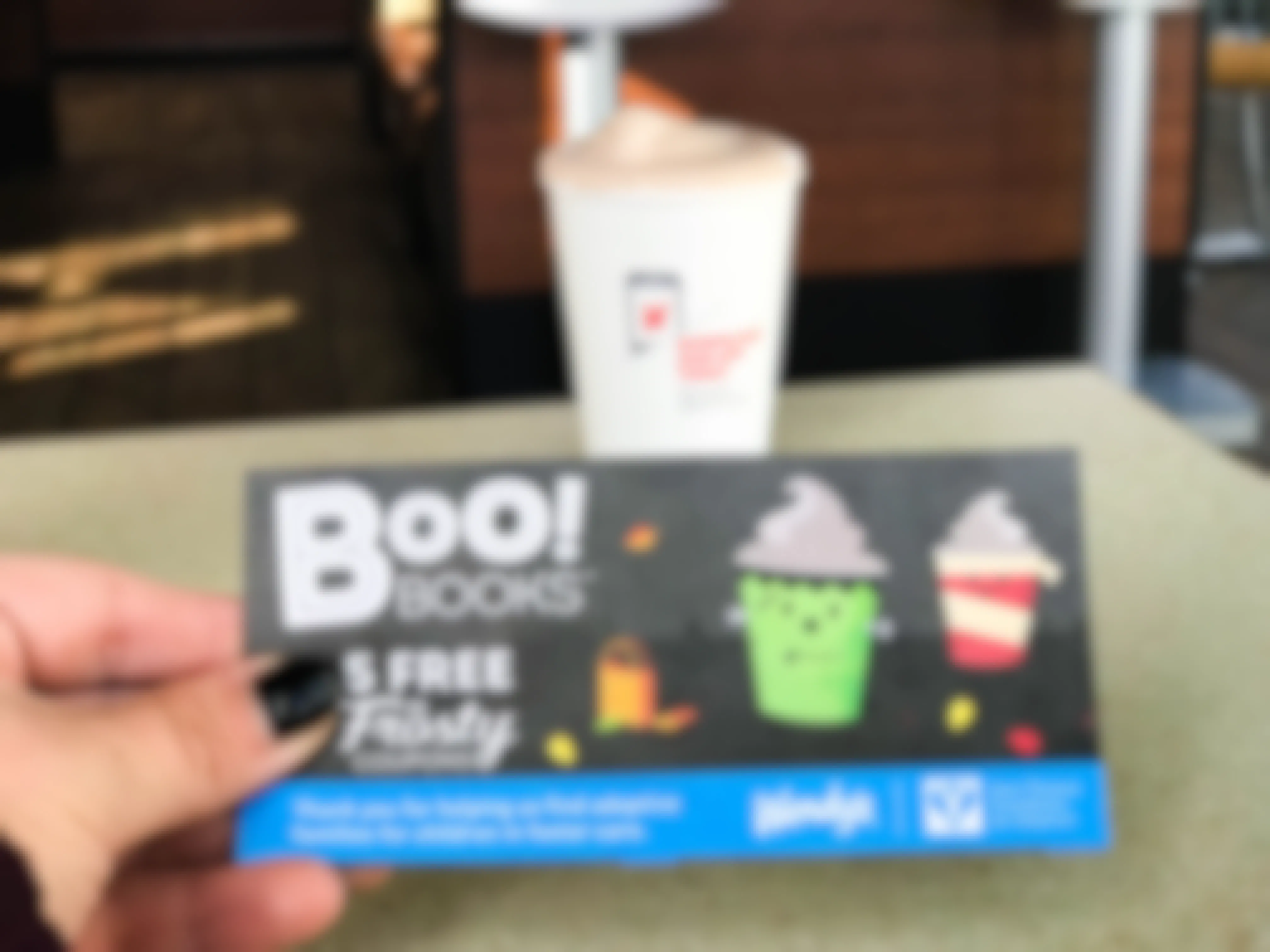 A person's hand holding a card that says, "Boo books 5 free jr frosty coupons" in front of a frosty on a table at Wendy's.