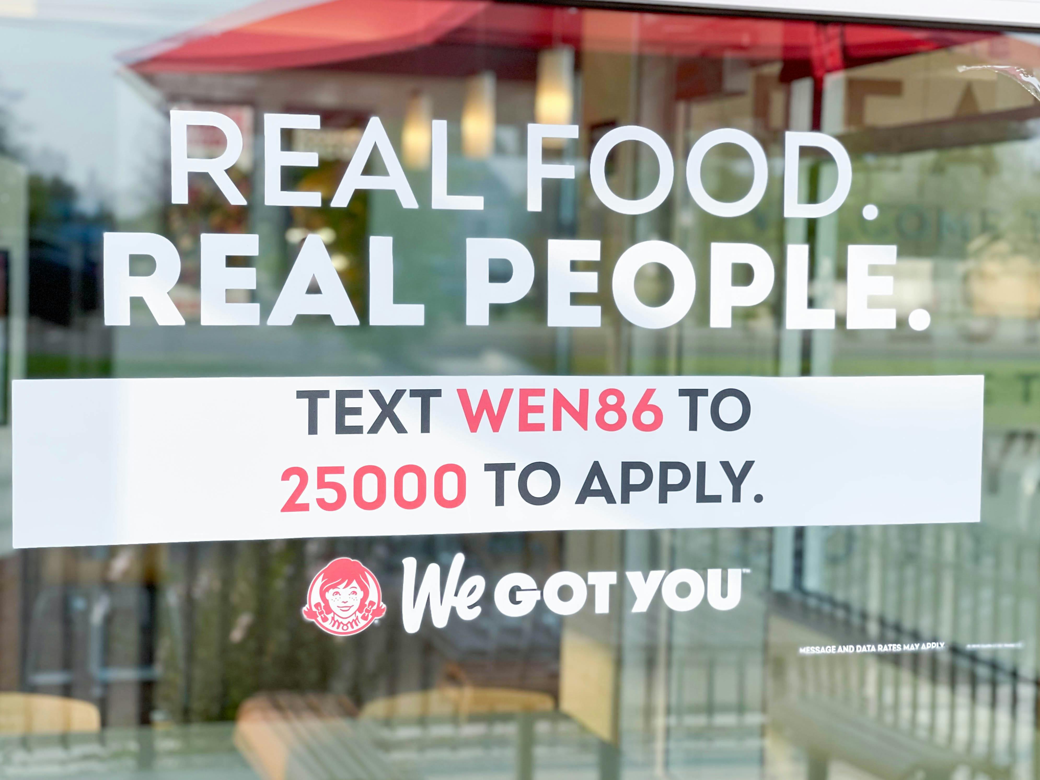A window cling in a Wendy's window that reads, "Real Food, Real People. Text WEN86 to 25000 to apply. We got you.