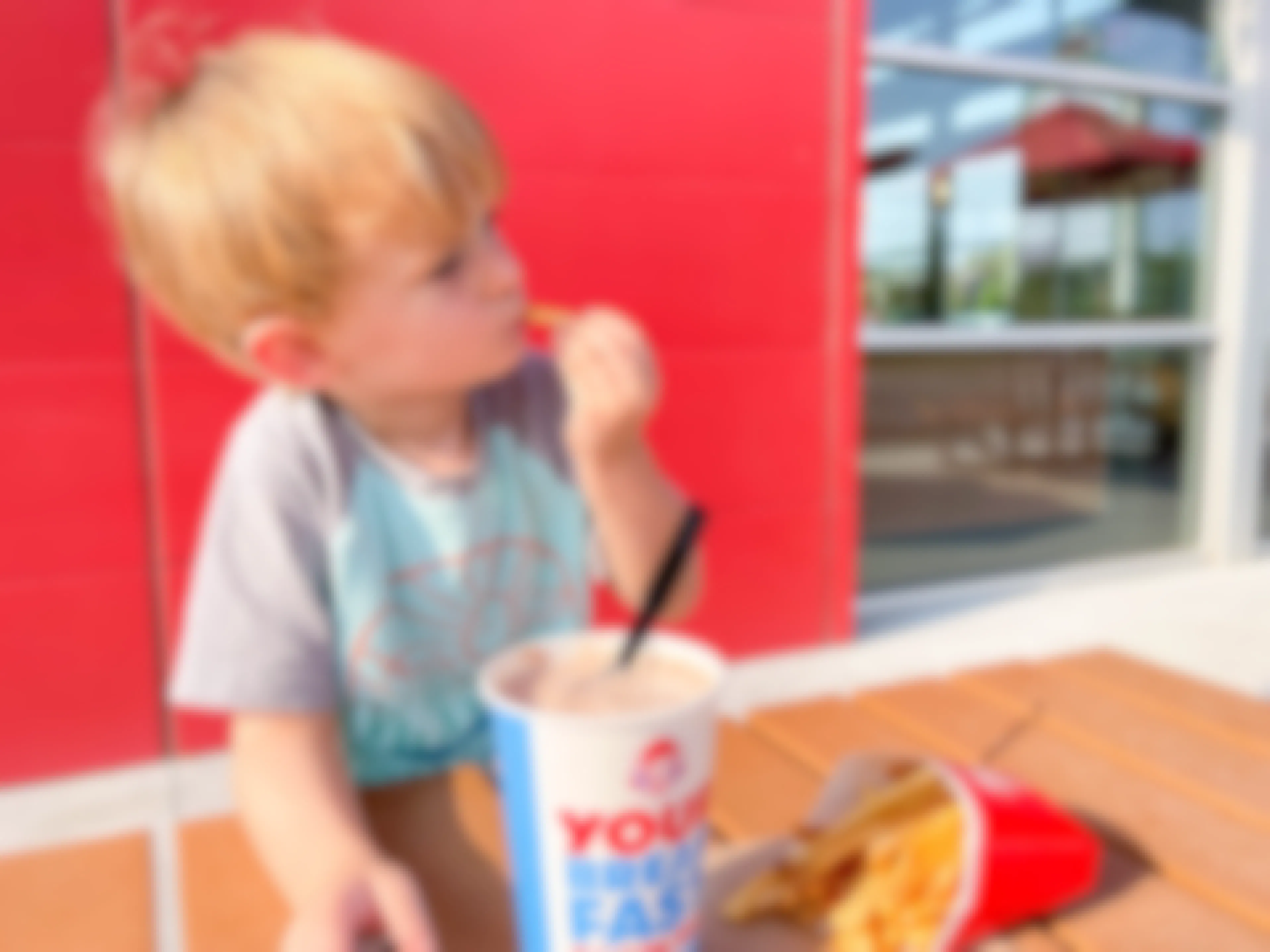 A child eating a french fry next to a box of Wendy's fries and a frosty at a table outside Wendy's.