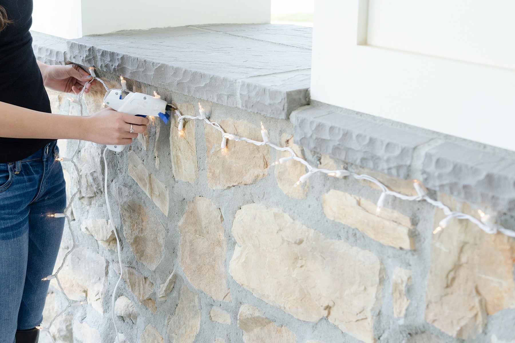 A person using a hot glue gun and glue to attach white Christmas lights to brick