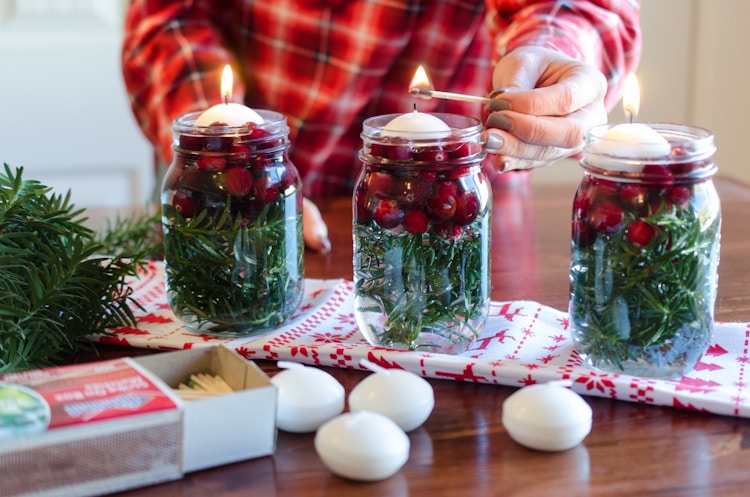 Floating candles being lit in mason jars filled with greenery and cranberries