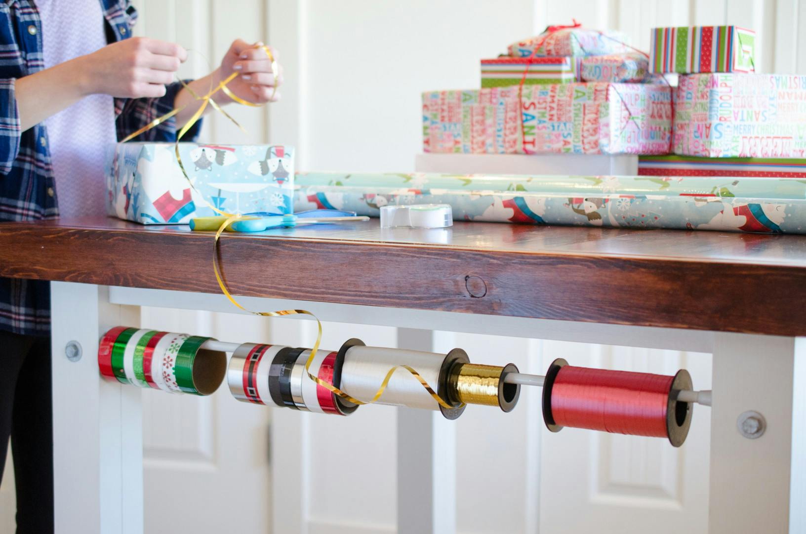 Gift wrap ribbon on a tension rod, attached under a kitchen table with wrapped packages and Christmas wrapping paper on top.